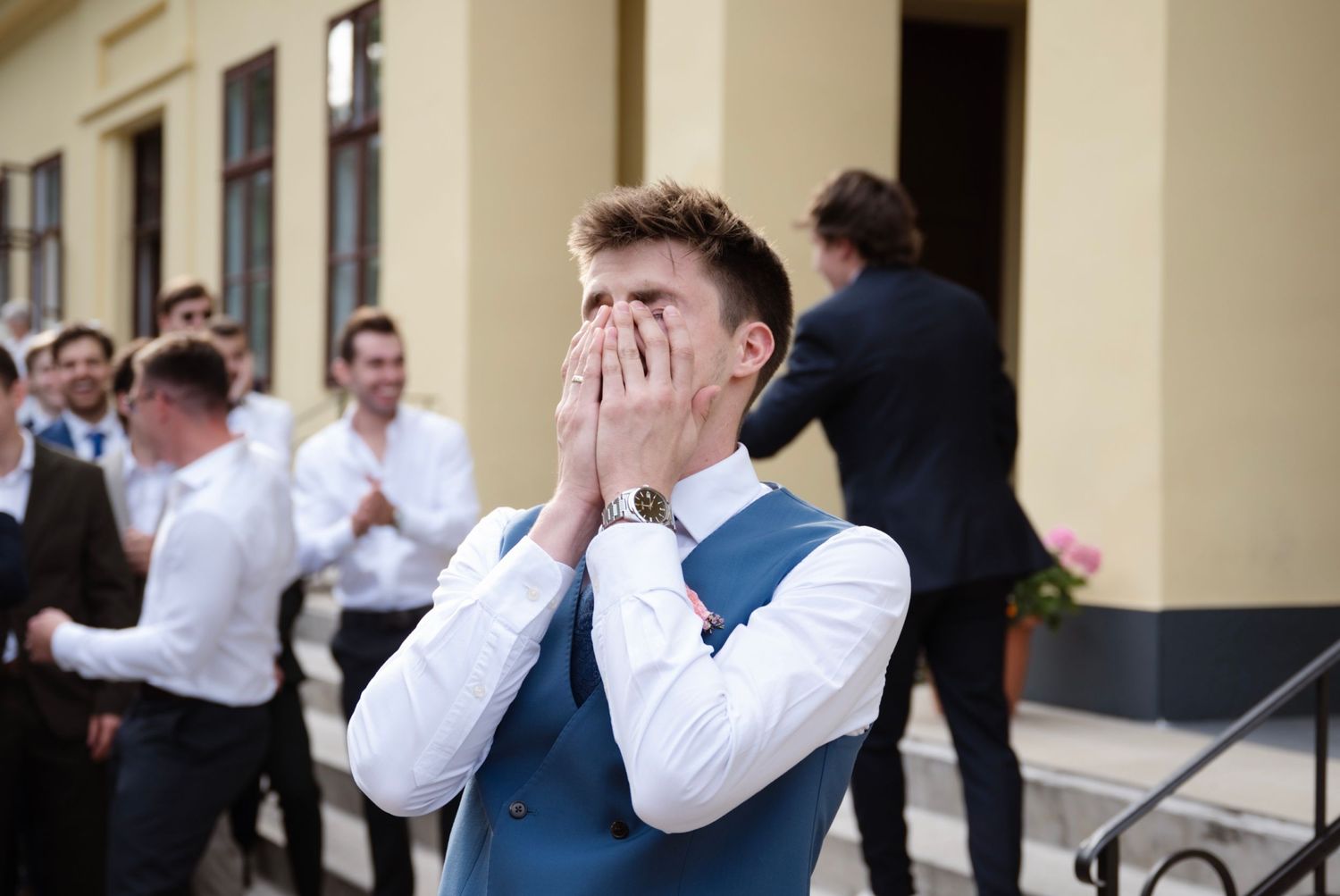 Groom covering his face with hands in emotional reaction, surrounded by groomsmen outside a wedding venue.