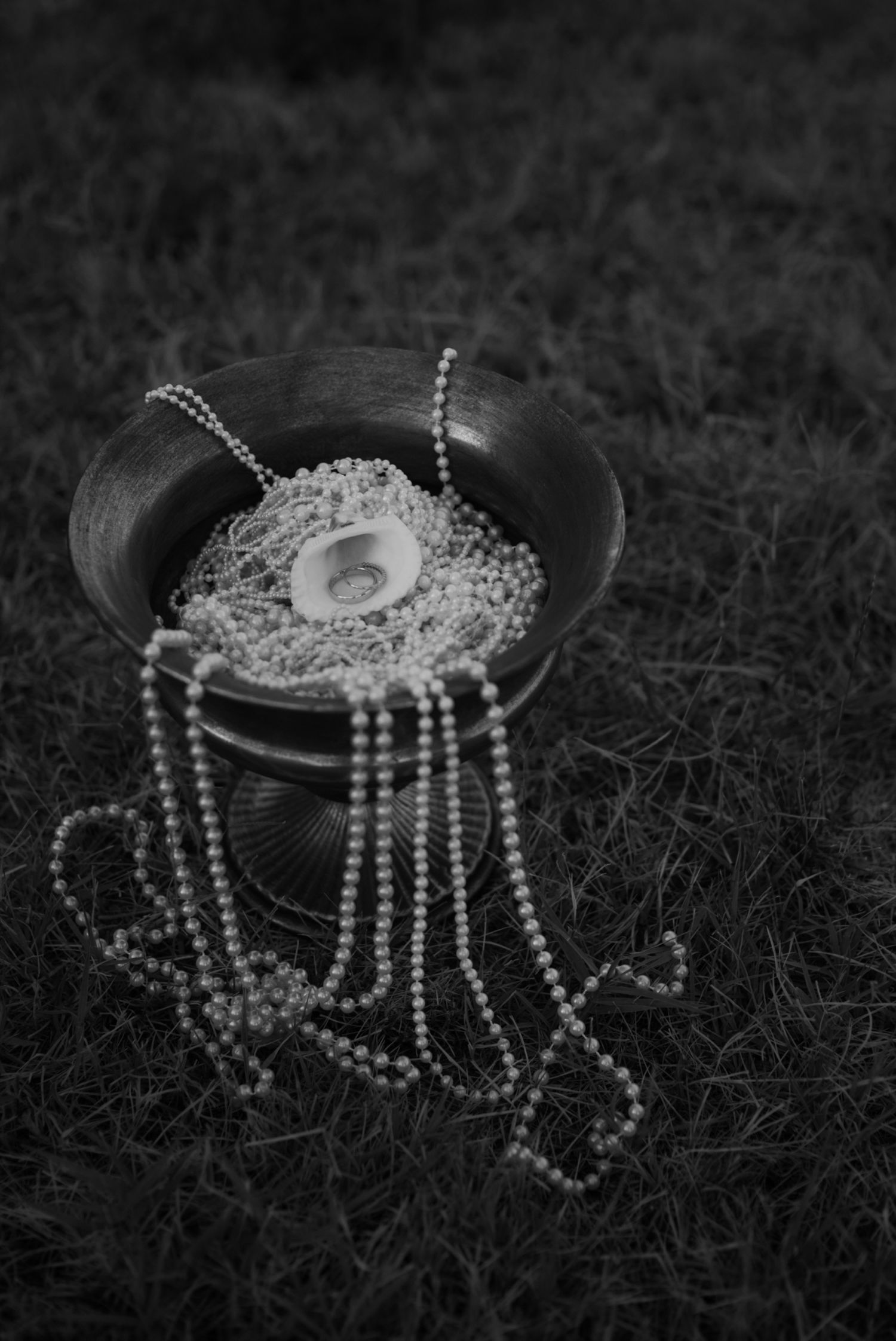 Black and white photo of pearl necklaces and a ring displayed in a decorative bowl on grass.