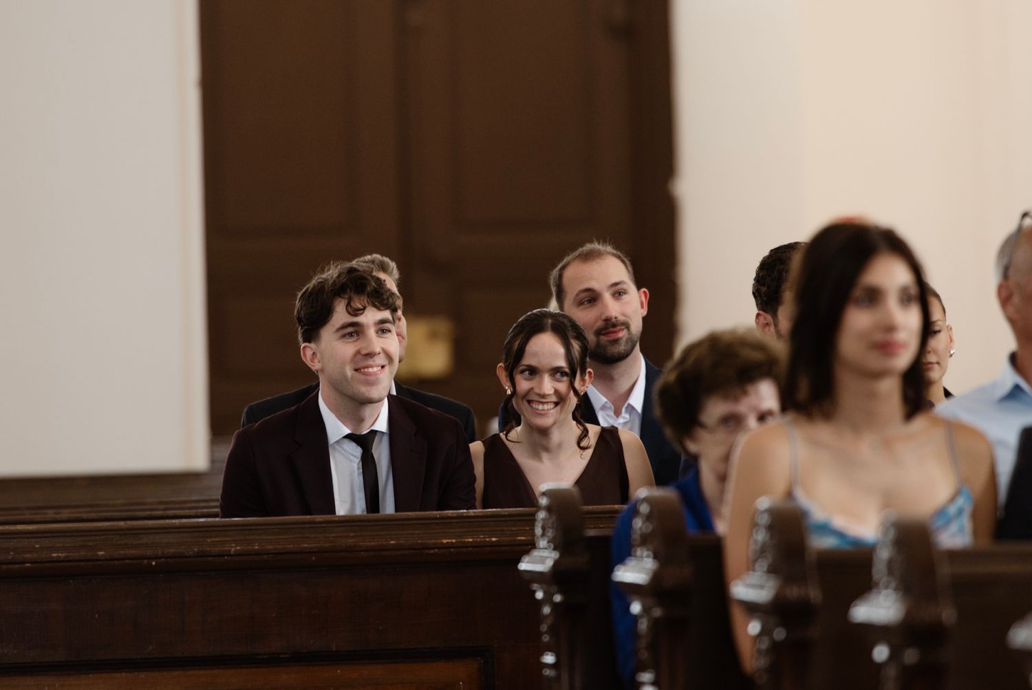 Guests smiling and seated in wooden church pews during a wedding ceremony indoors.