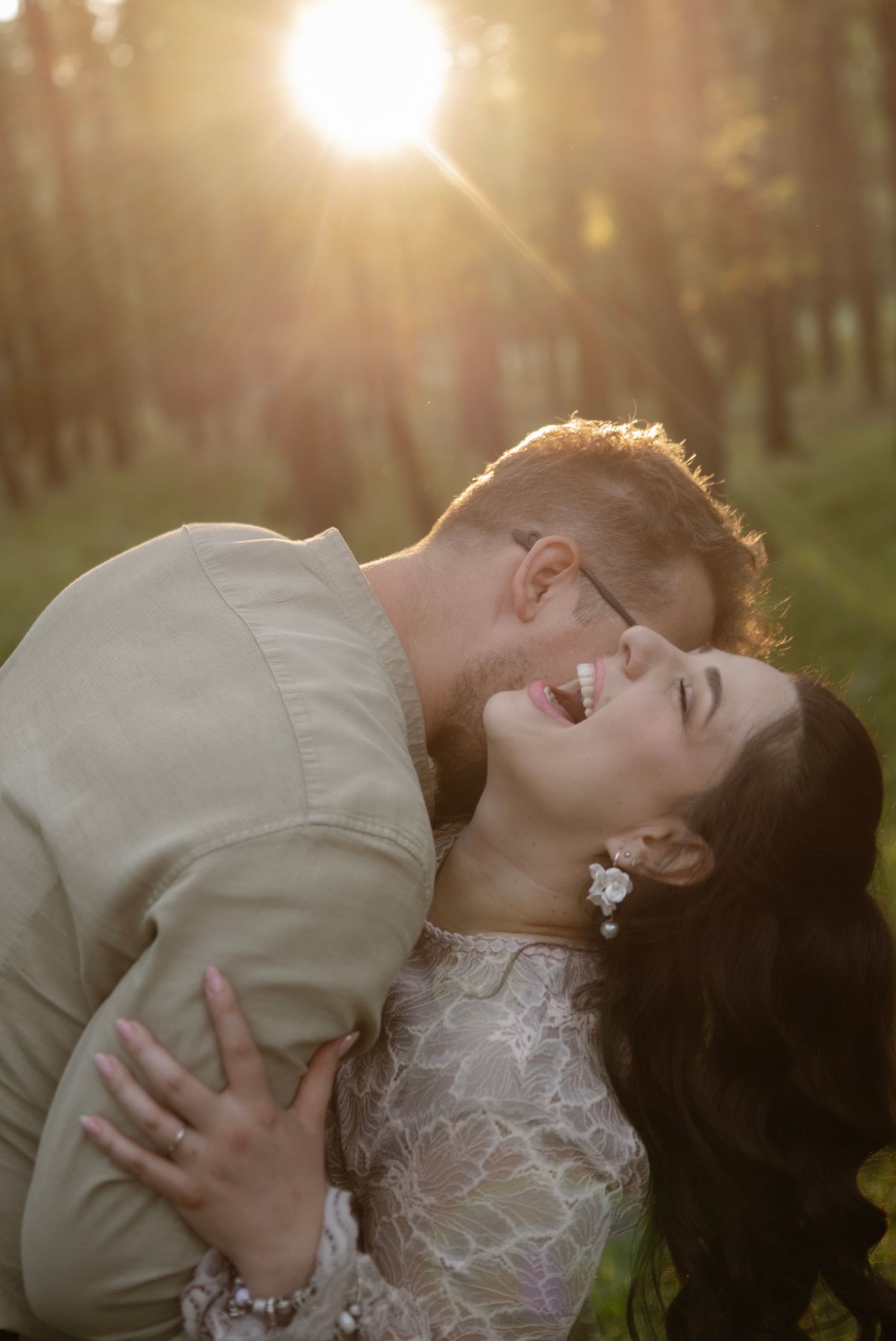 A joyful couple shares a romantic embrace in a sun-drenched forest, the bride laughing in a lace gown at golden hour.
