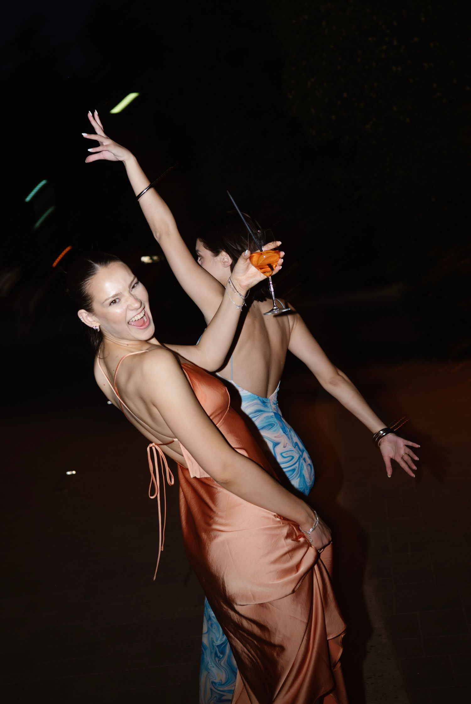 Woman in copper satin gown dances joyfully holding cocktail at outdoor evening wedding reception.