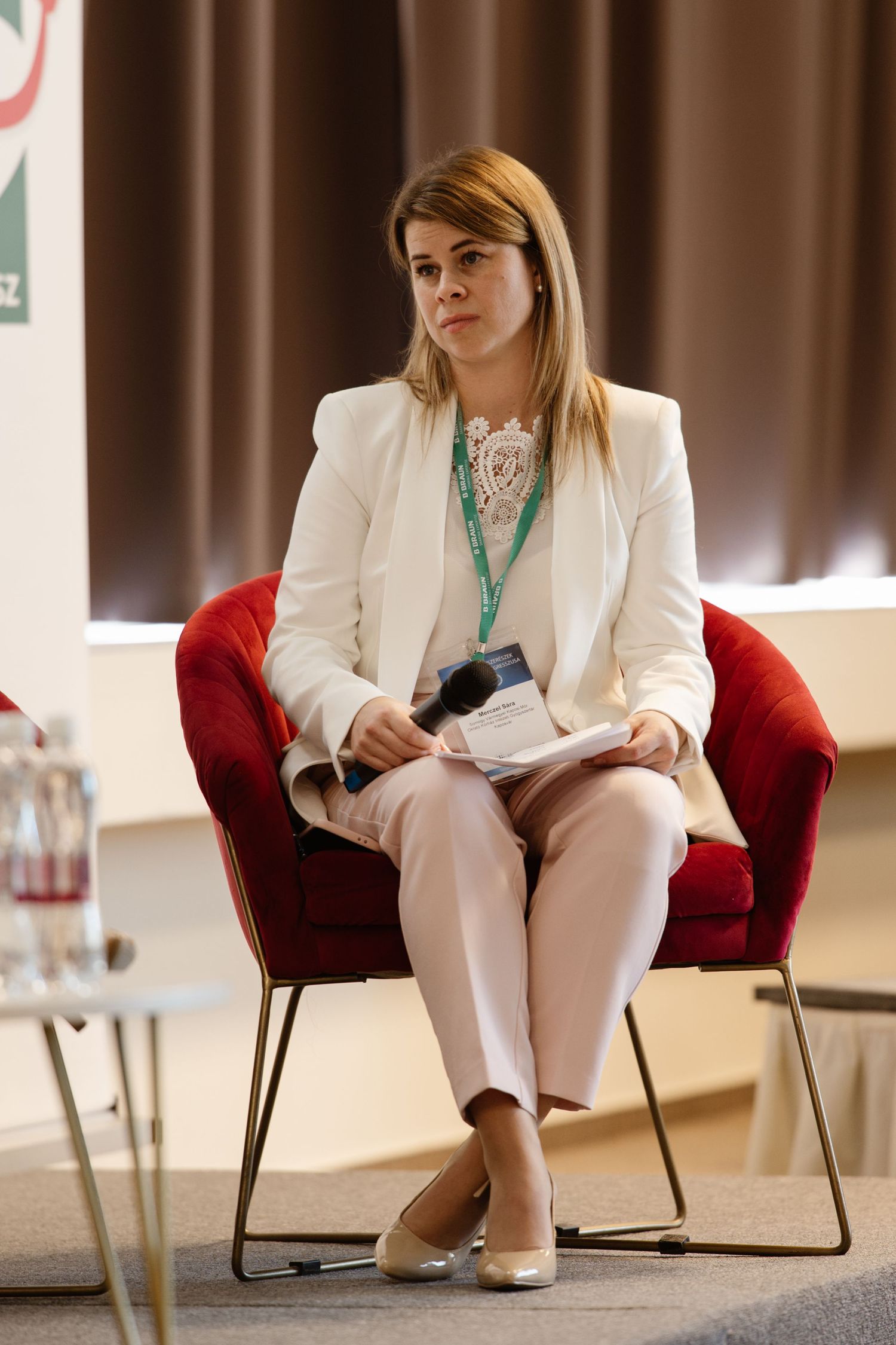 Professional wearing a white blazer sits in a red velvet chair during a conference presentation.