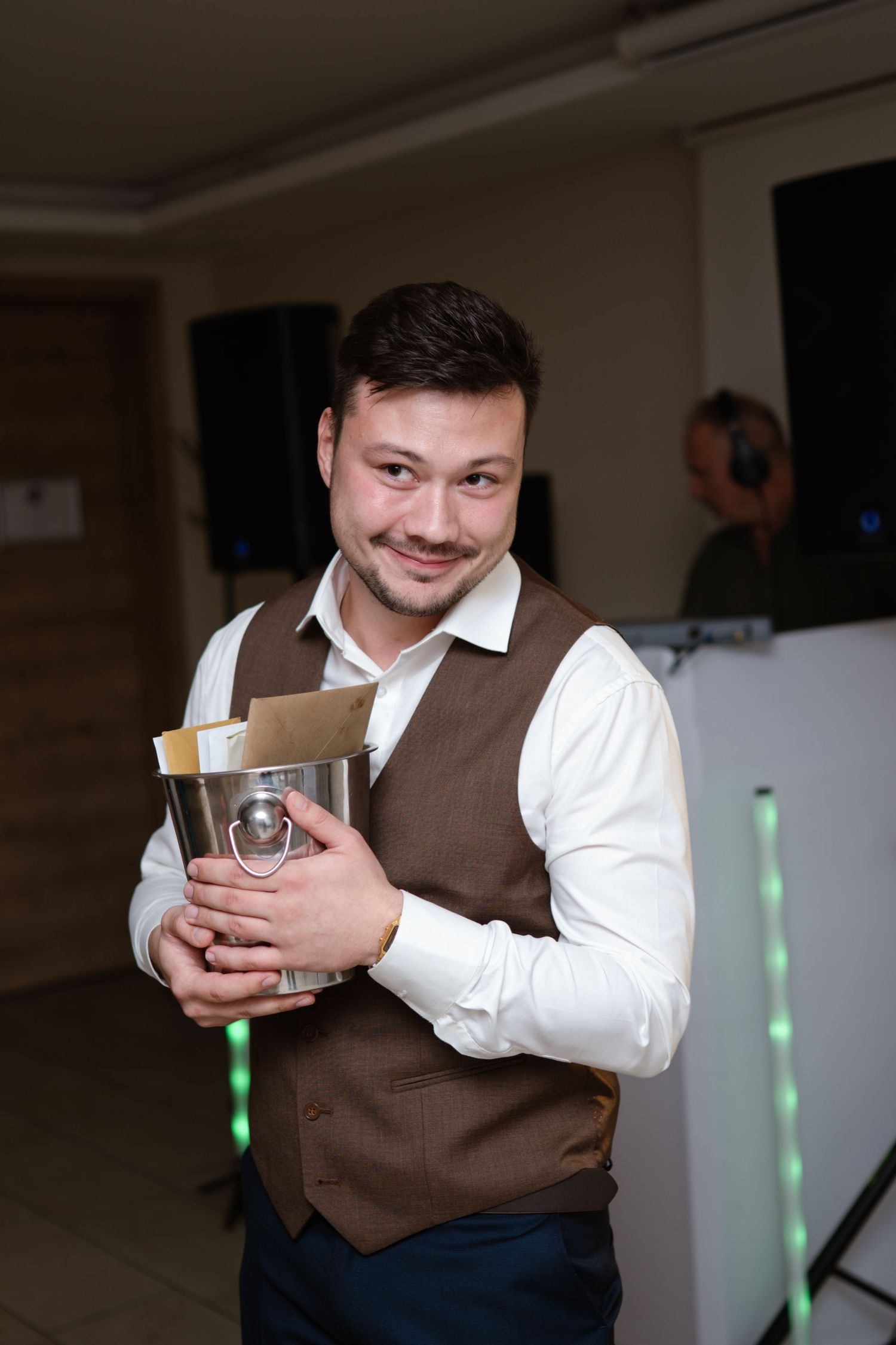 Person in brown vest and white shirt smiling while holding a drink at event.