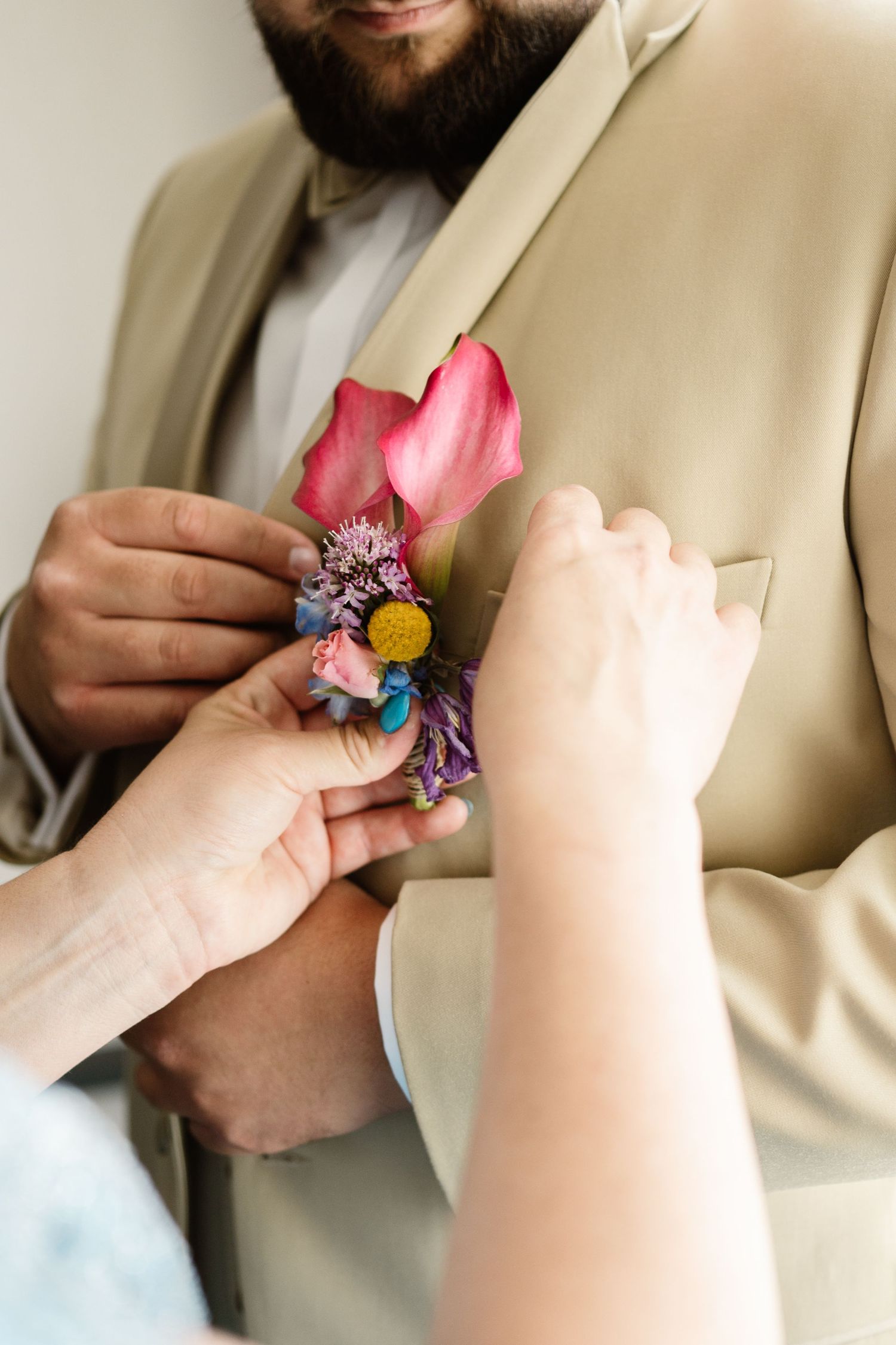 Close-up of hands pinning a colorful floral boutonniere to a tan suit jacket lapel.