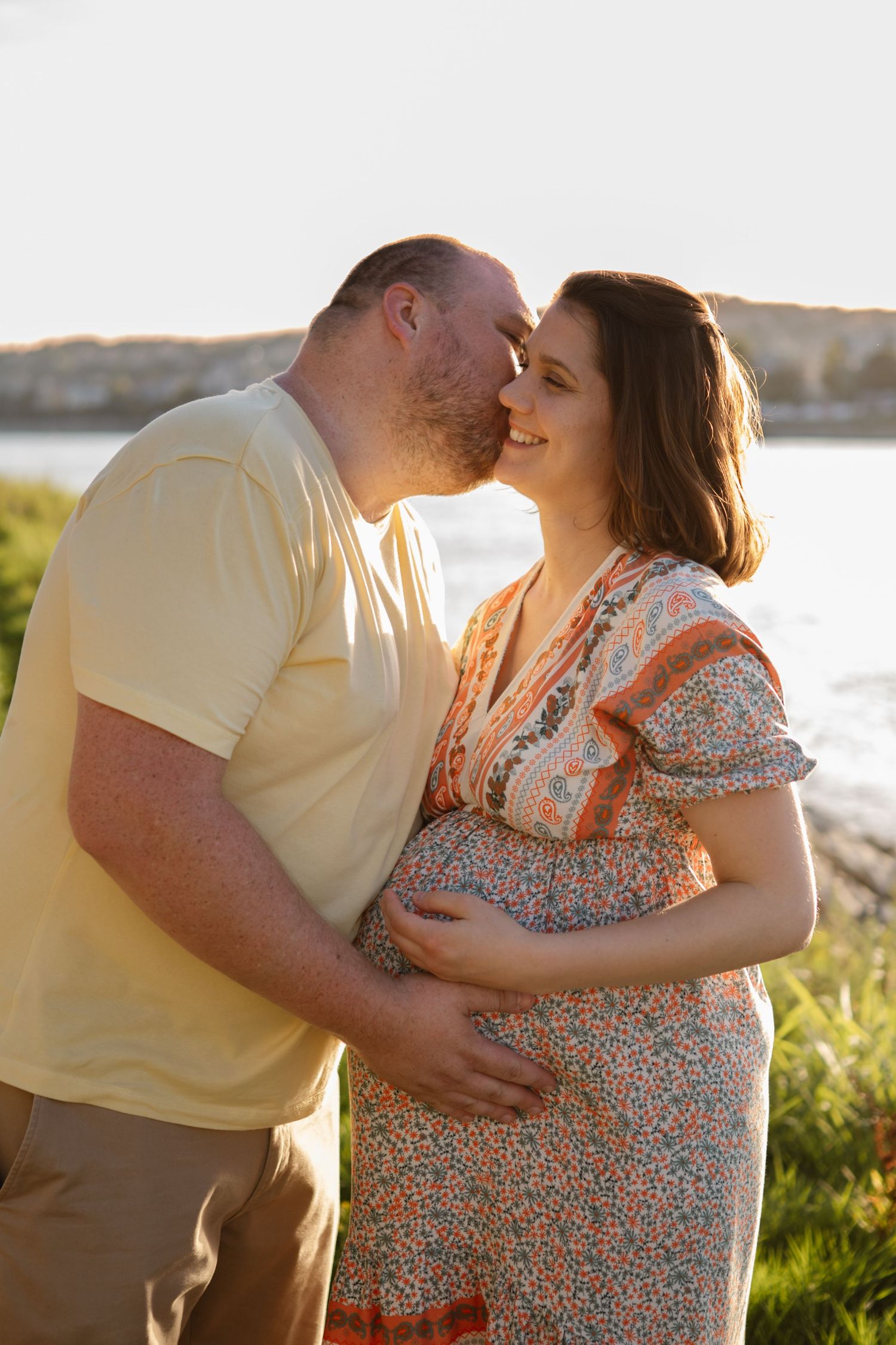 An intimate maternity photo of a couple embracing by the water during sunset.