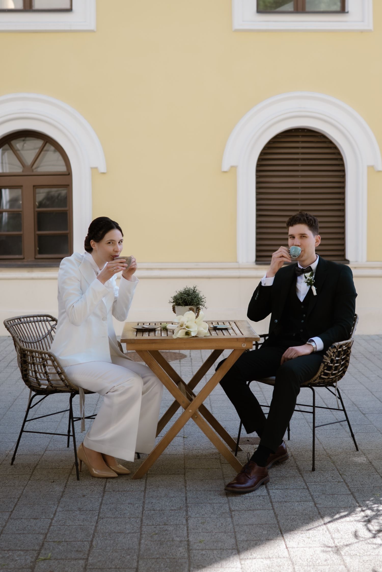 A bride and groom sit at an outdoor cafe table sipping coffee in front of a yellow European building.