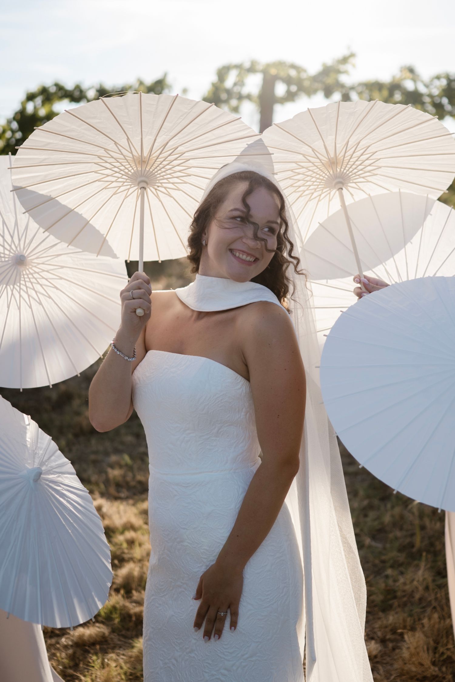 Smiling bride in white strapless gown holding white parasol outdoors, surrounded by multiple white paper umbrellas.