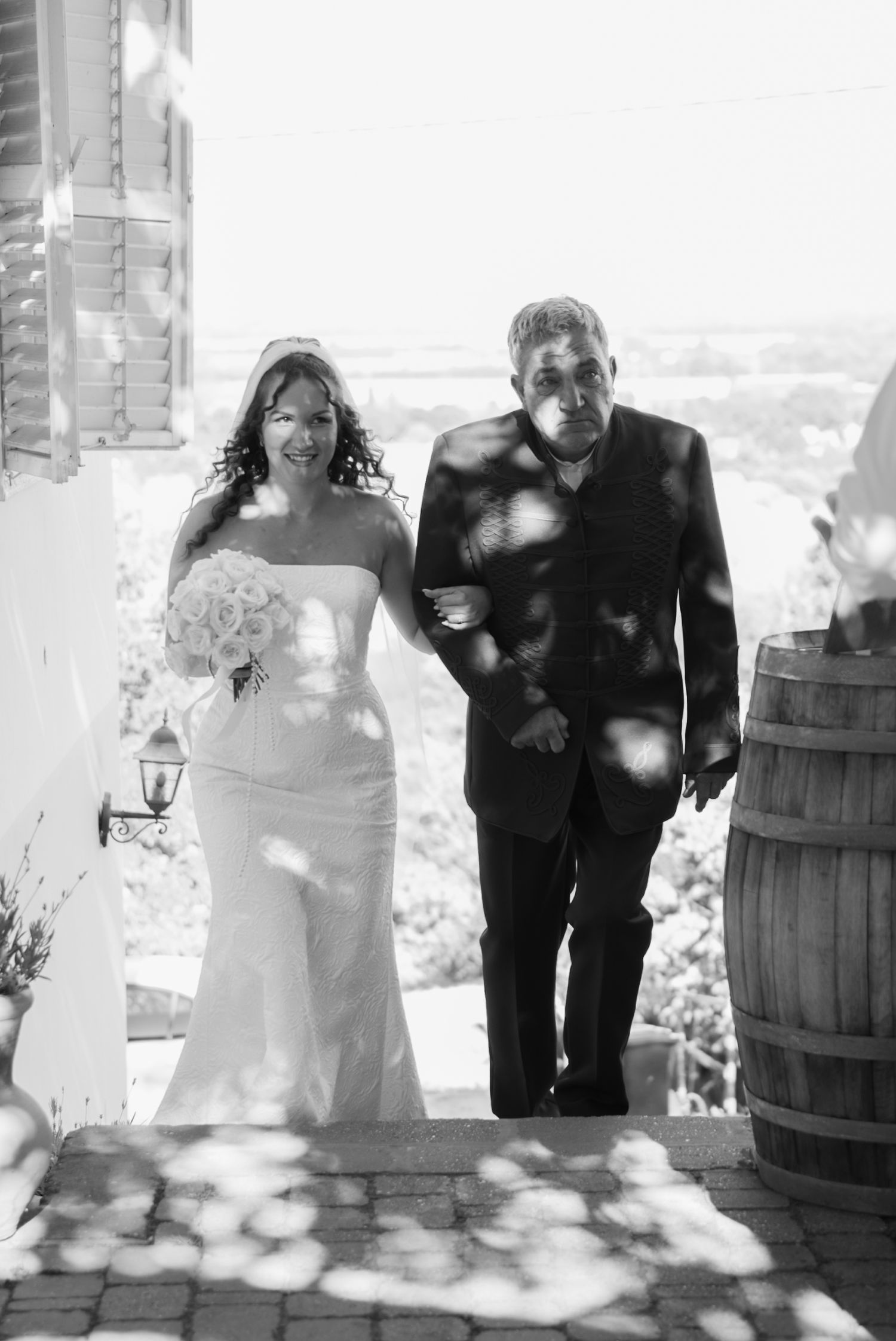 Black and white photo of bride in strapless gown walking with escort at outdoor wedding.