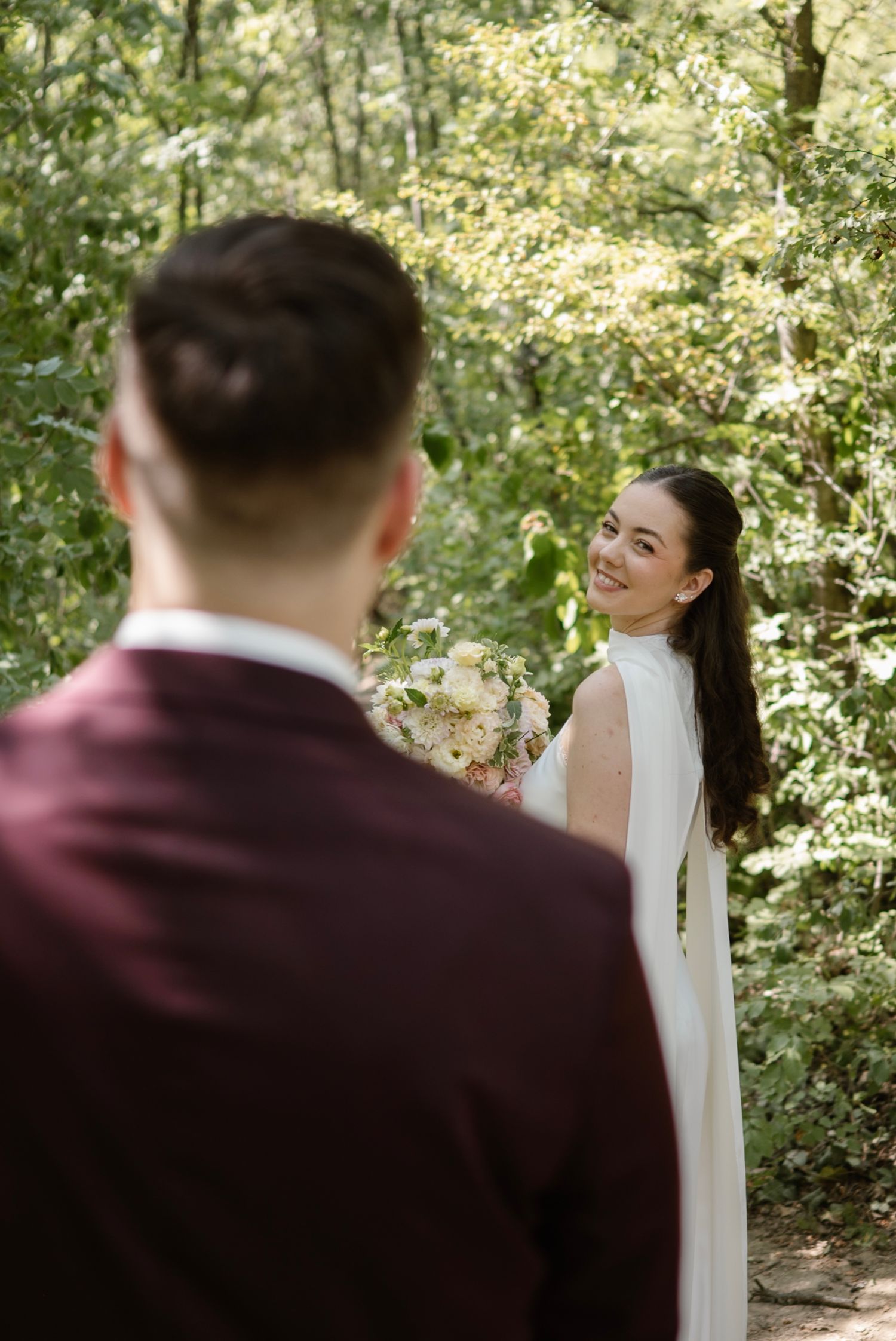 Bride in white cape gown smiles at groom in burgundy suit during outdoor forest wedding first look.