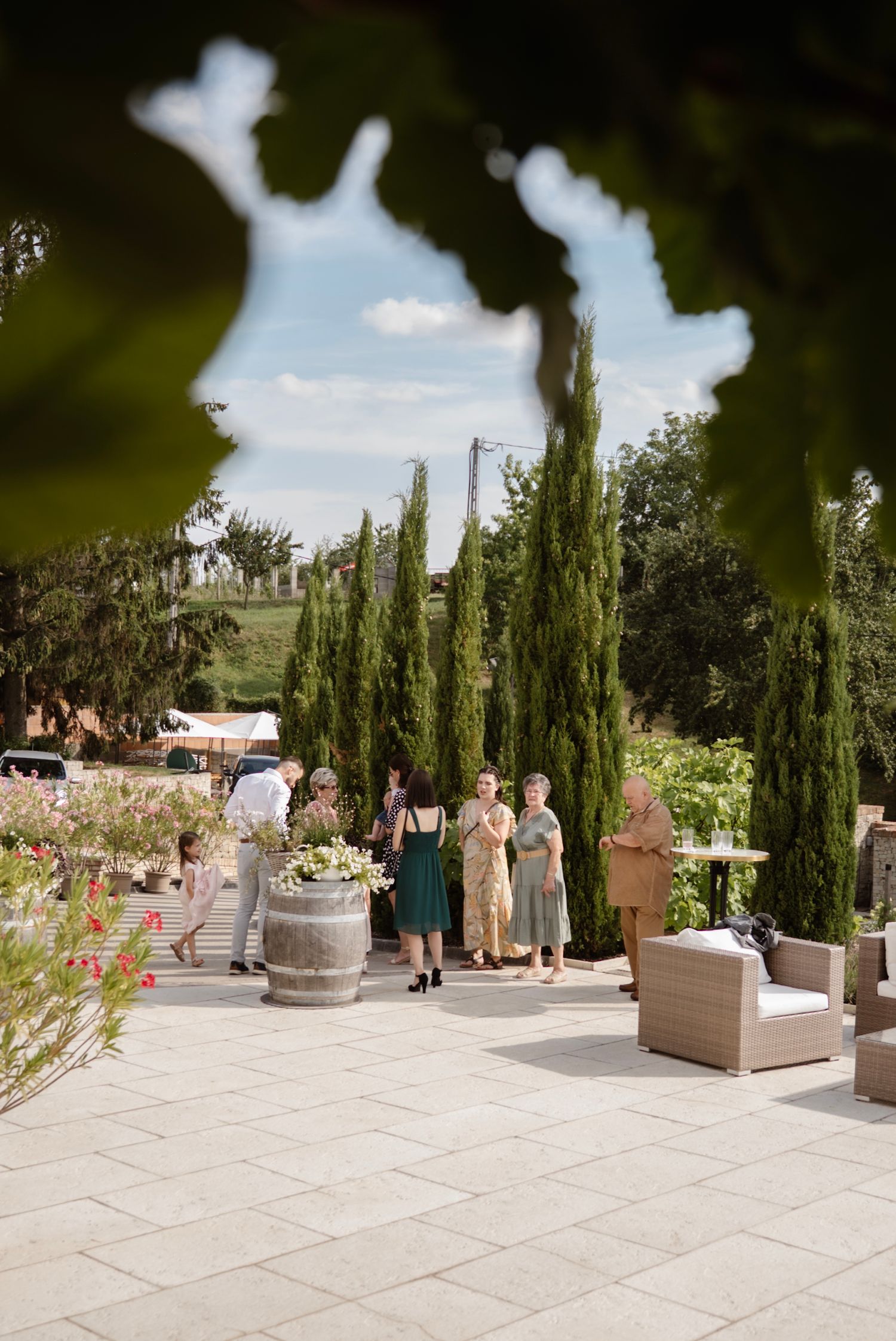 Guests mingle on a stone patio surrounded by tall cypress trees and flower gardens at an elegant outdoor venue.