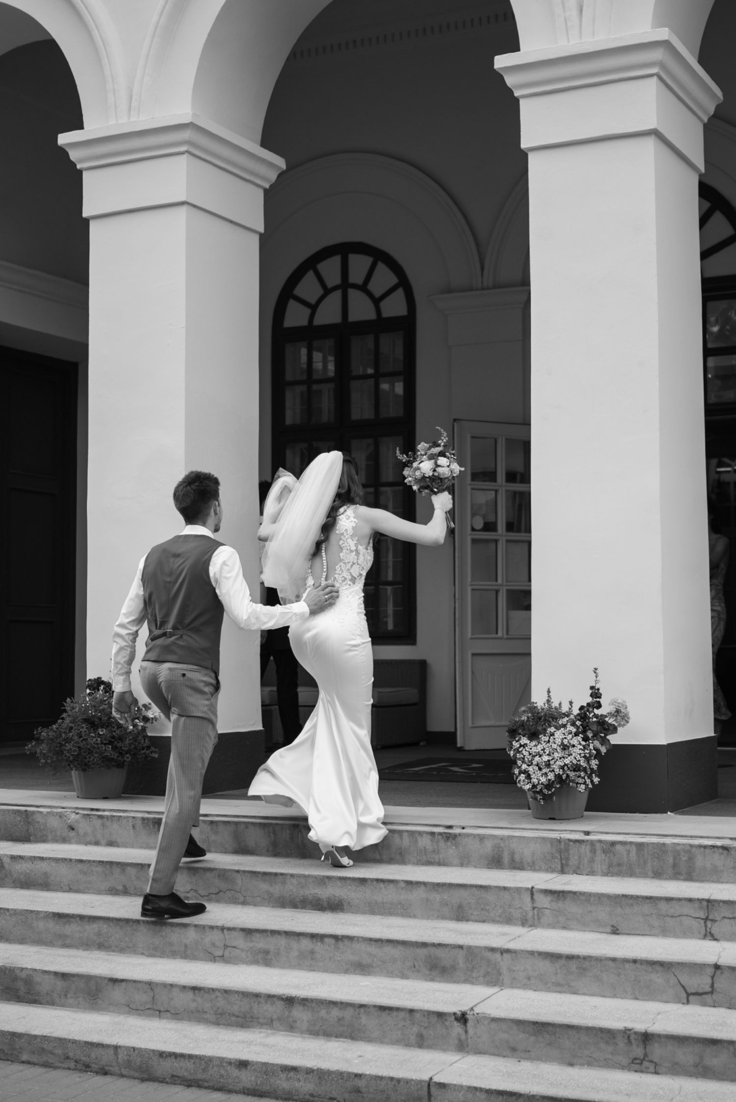 Black and white photo of a bride and groom walking up steps toward elegant building entrance on their wedding day.
