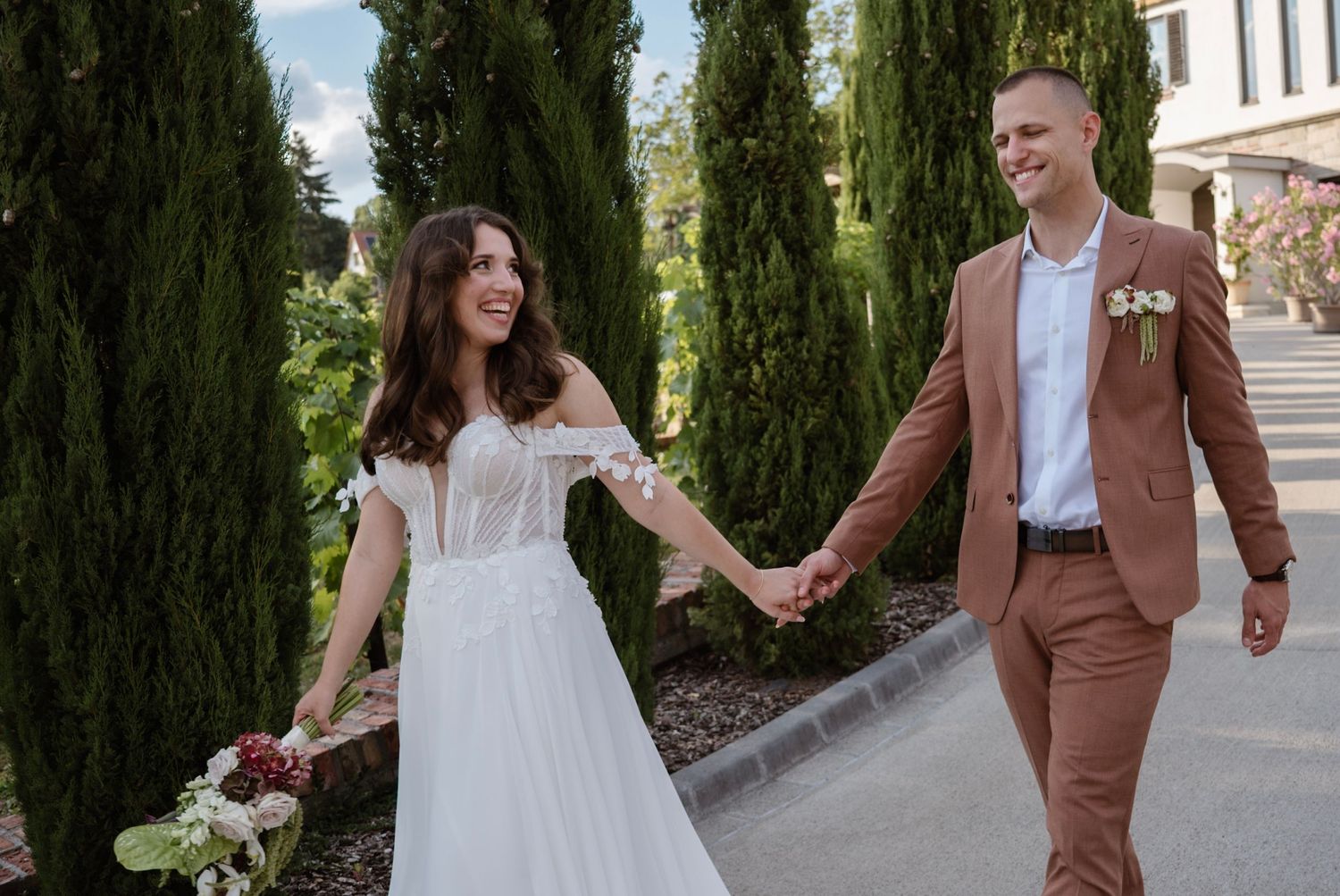 Bride in white gown and groom in tan suit hold hands walking among tall cypress trees at outdoor wedding venue.