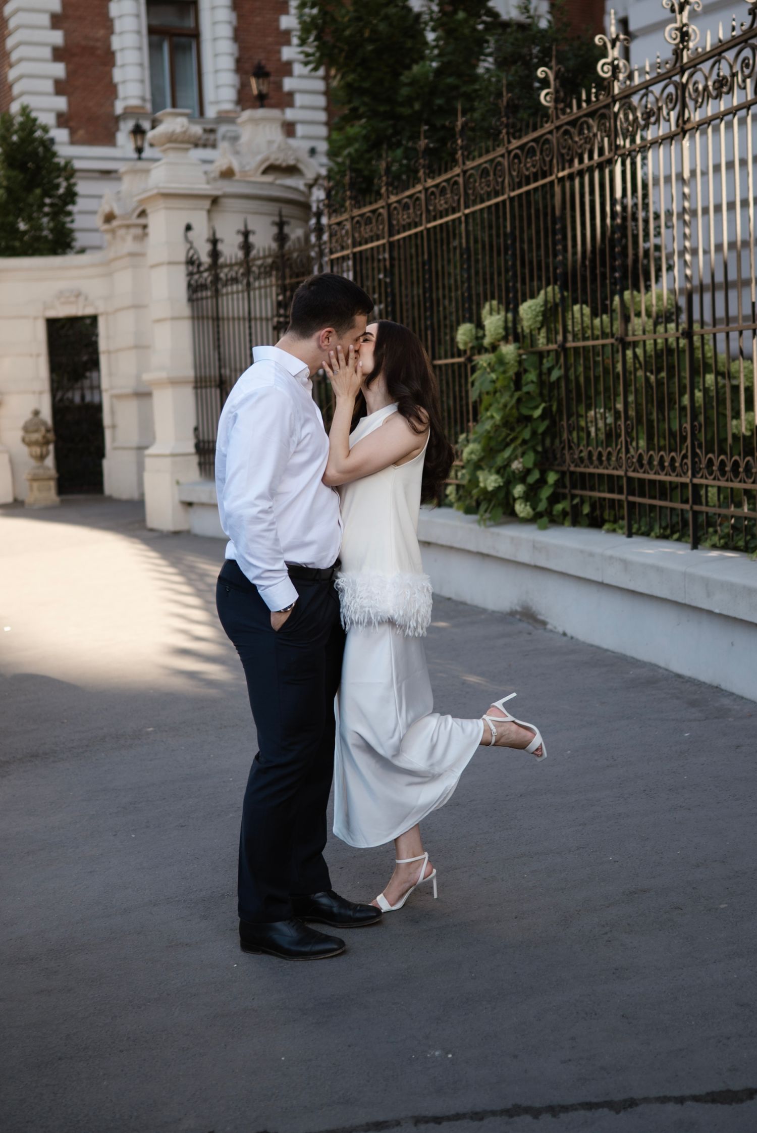 Couple sharing a romantic kiss outdoors near ornate iron fence, woman in white feathered dress lifting her heel.