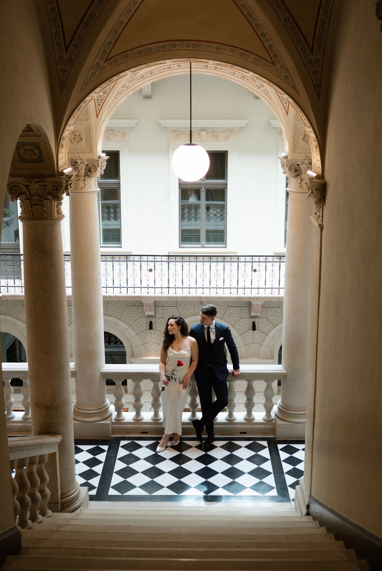 Bride and groom walk through elegant arched hallway with checkered floors and ornate columns in a grand historic building.