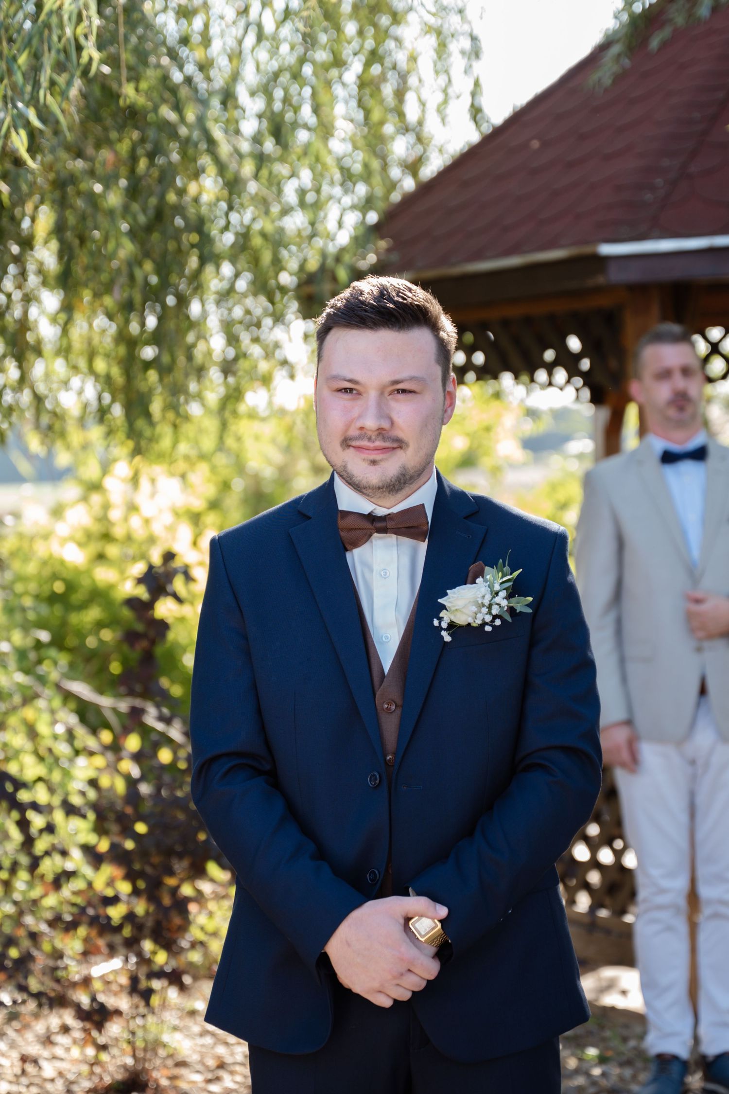 Groom in navy suit with boutonniere standing in front of outdoor ceremony space.