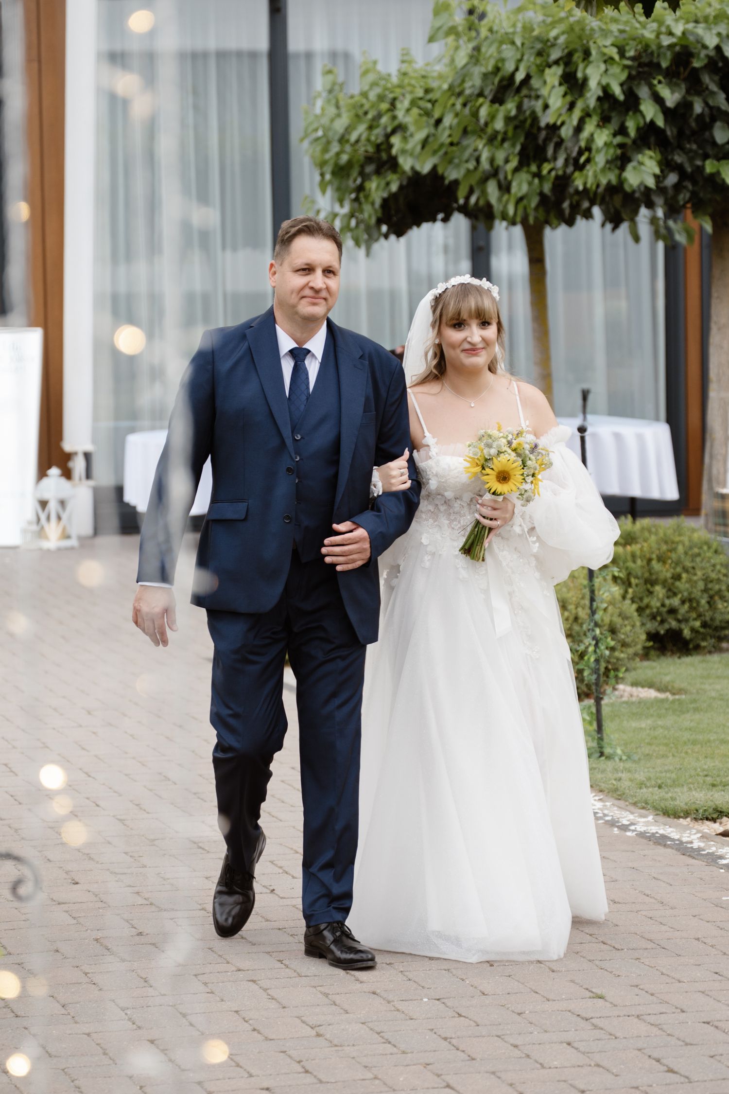 A couple walking down a brick pathway with white floral decorations and string lights.