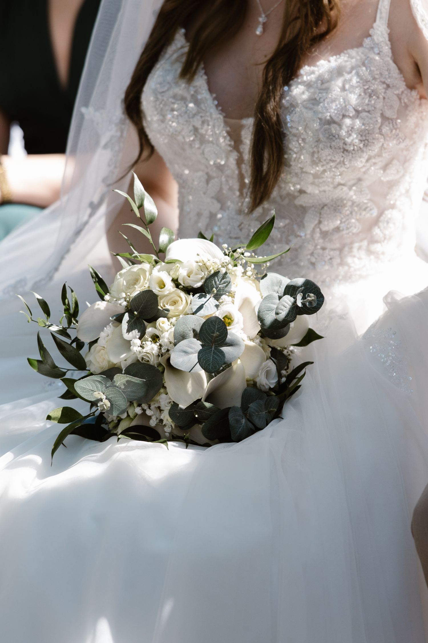 Close-up of an elegant white bridal bouquet with eucalyptus and roses against a lace wedding dress.