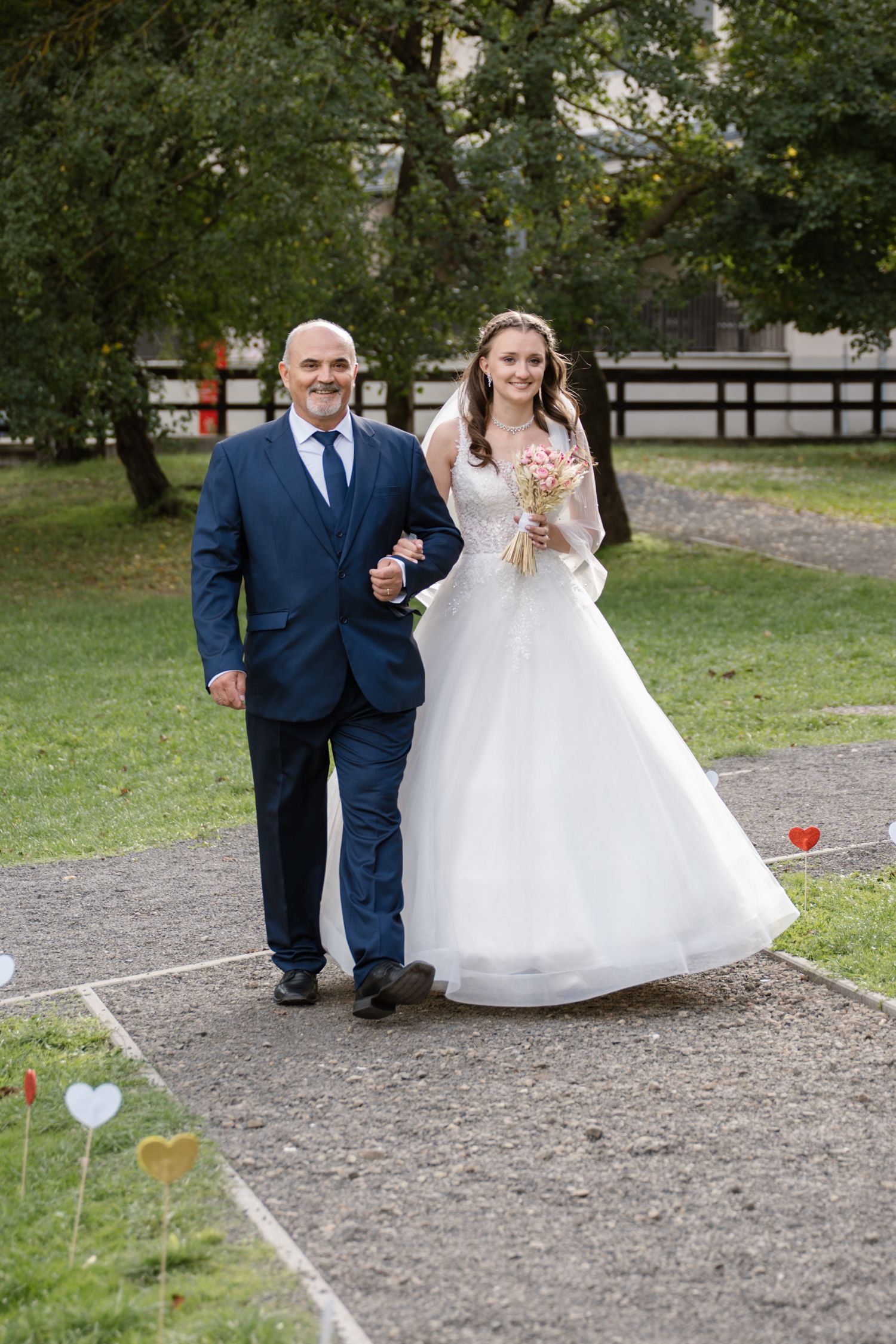 A wedding processional on a garden path surrounded by green trees and grass.