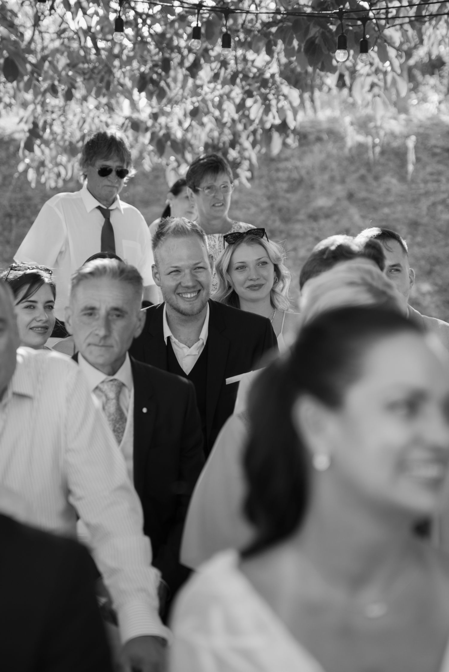 Black and white photo of smiling wedding guests seated outdoors during a ceremony.