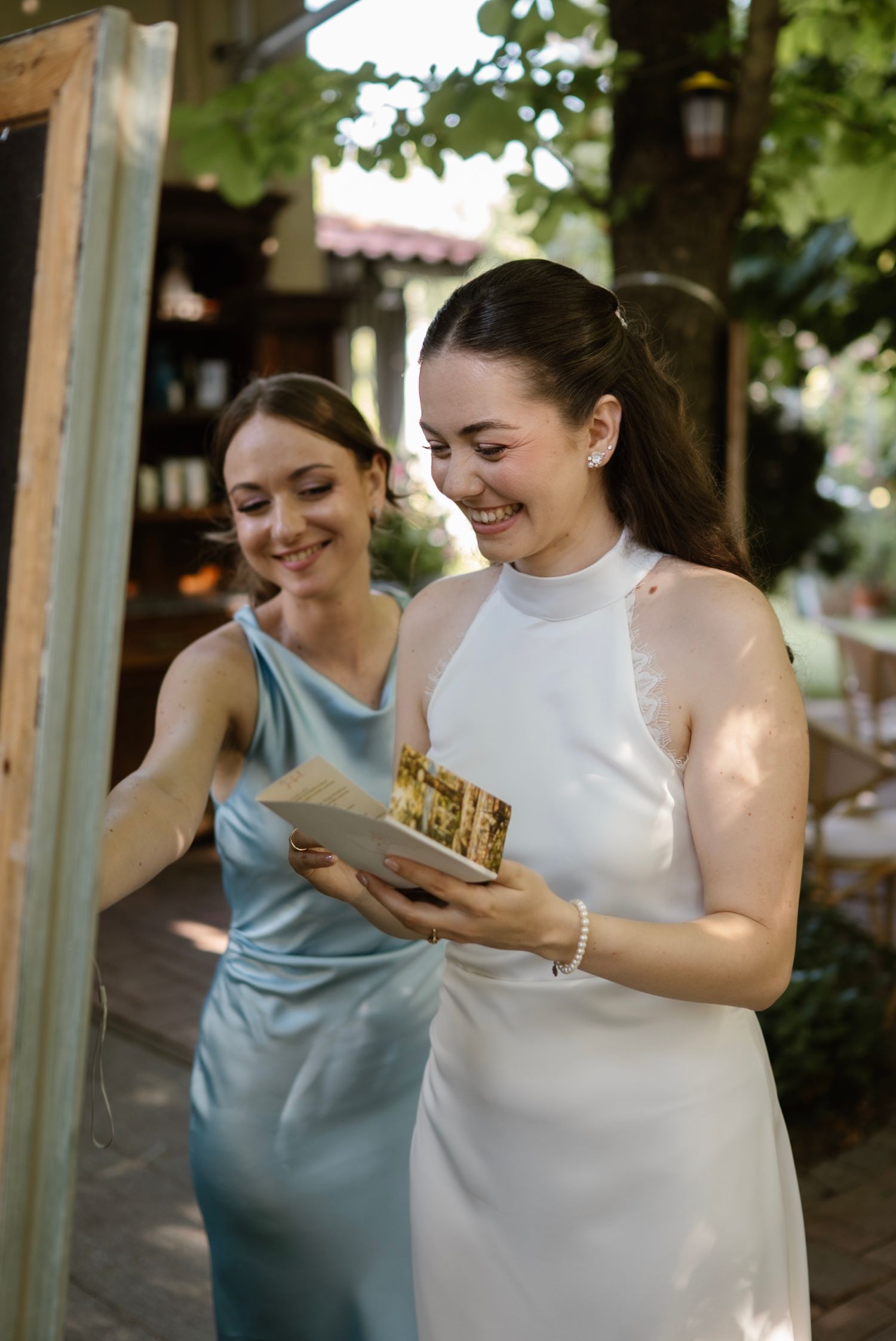 Bride in white halter dress smiles while holding a gift box with bridesmaid in blue satin dress outdoors.