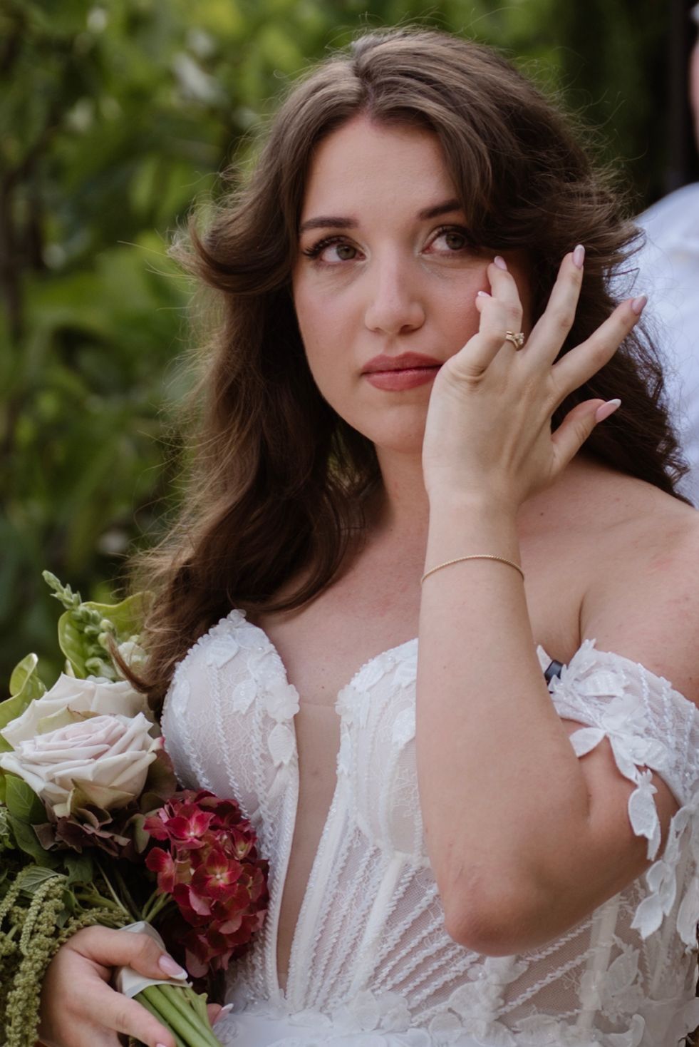 A bride in a white lace gown holds a bouquet of roses, gazing upward with a dreamy expression outdoors.