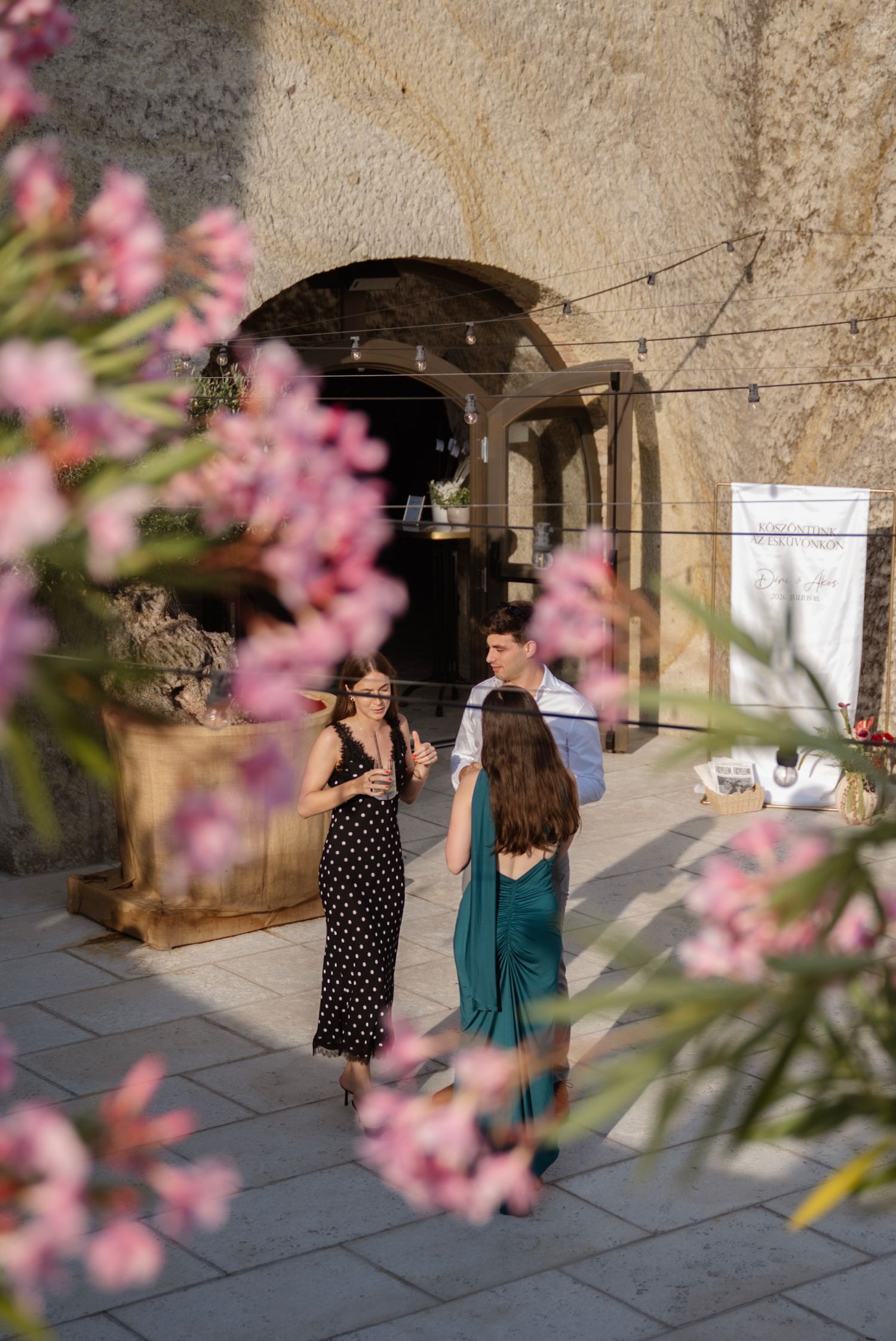 Guests in elegant attire socialize outdoors at a rustic venue, framed by pink flowering branches.