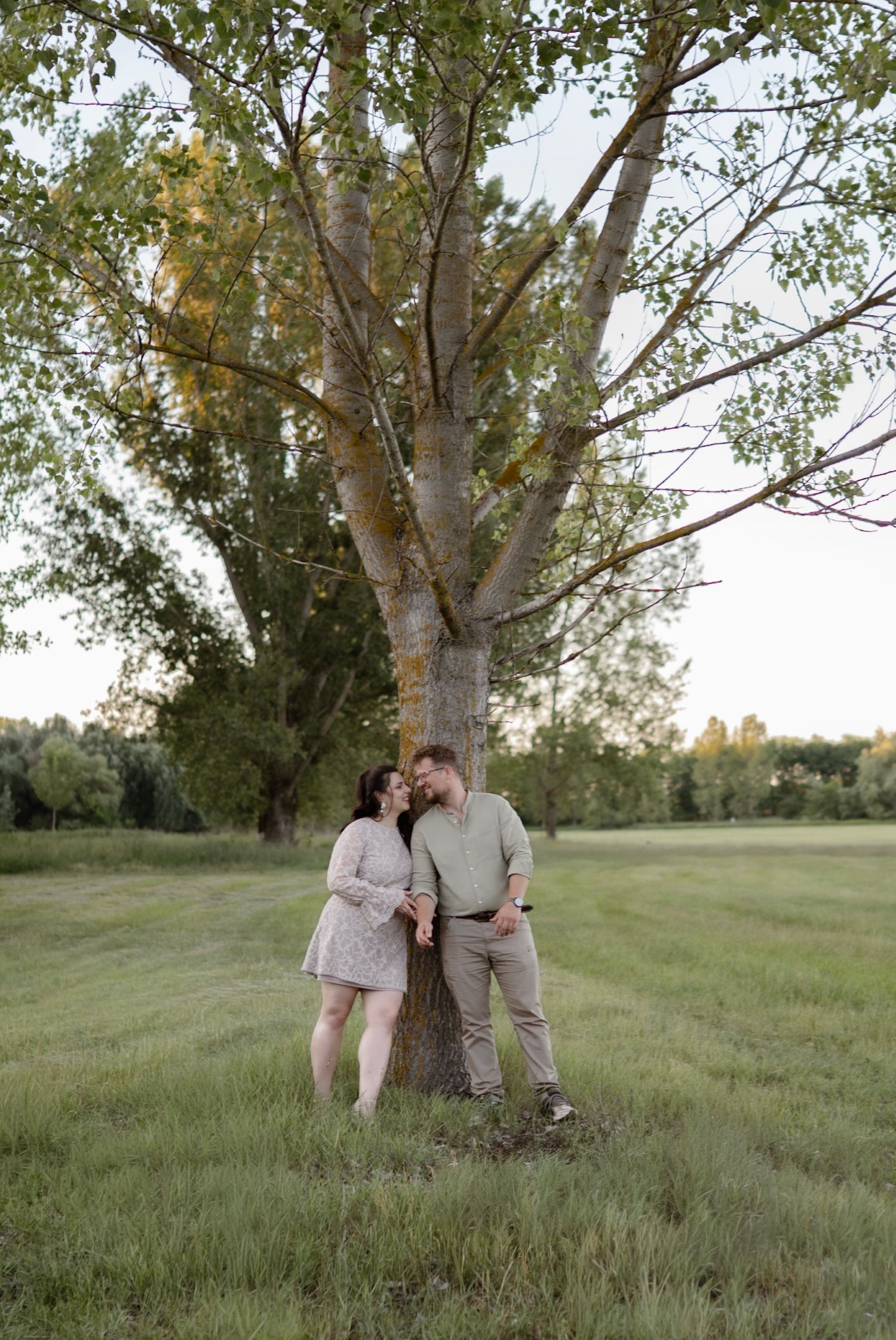 Couple leaning against a large tree in a green field during golden hour engagement photo session.