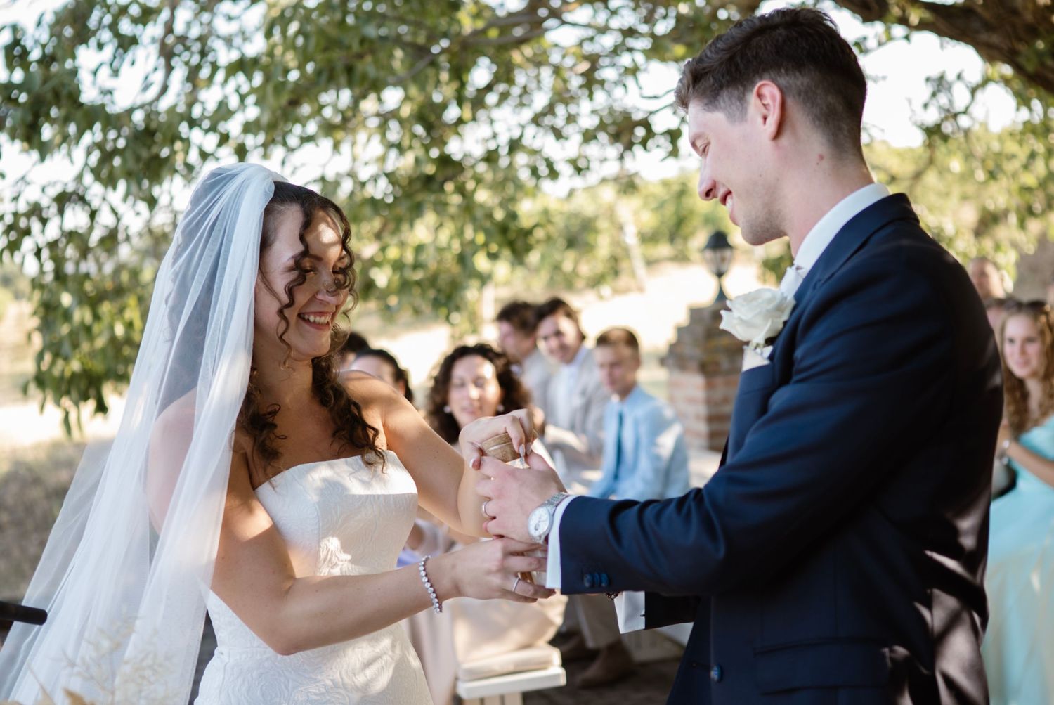 Bride and groom exchanging rings during outdoor wedding ceremony with guests seated in background.