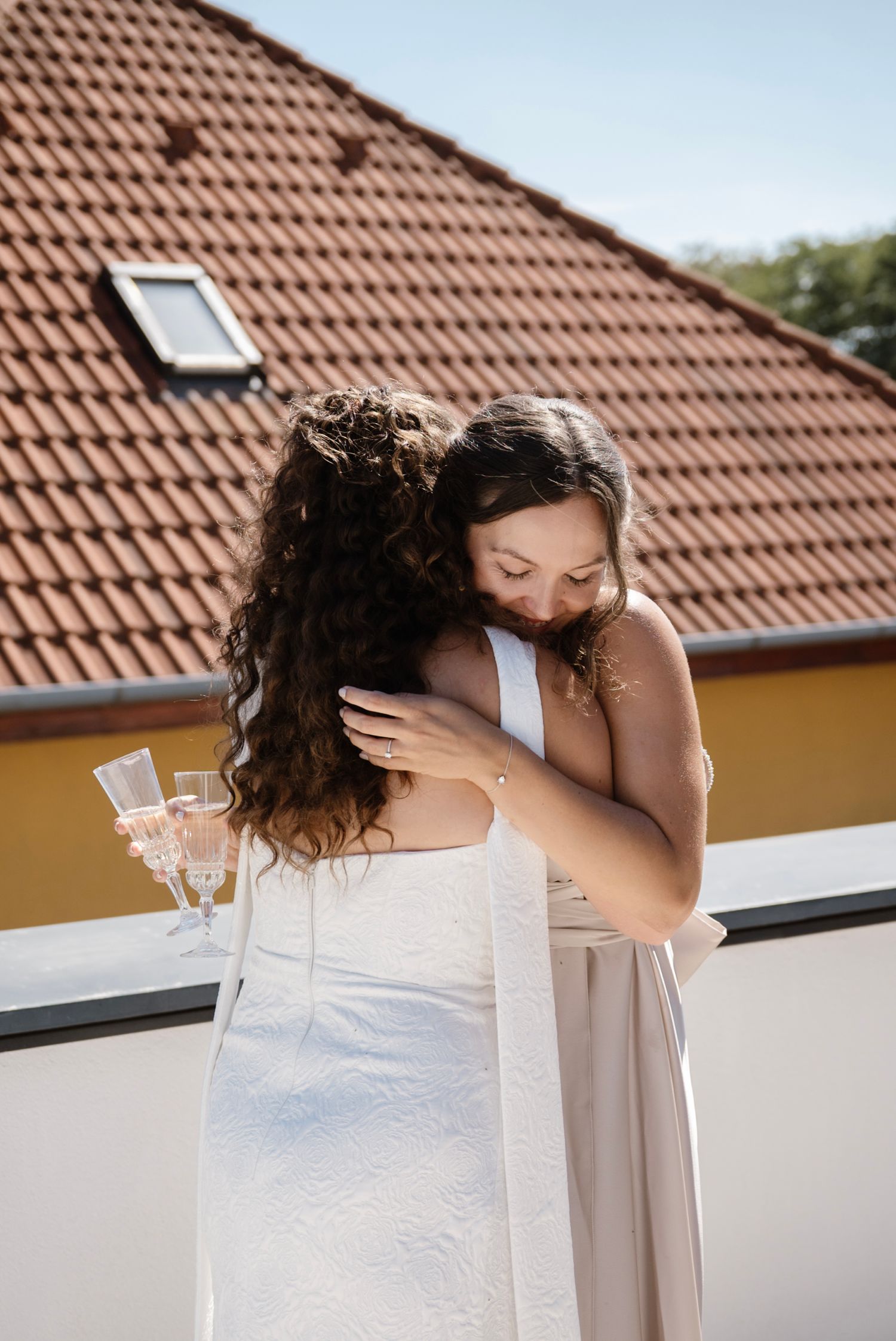 Bride in white wedding dress sharing a warm hug with bridesmaid on sunny outdoor balcony.