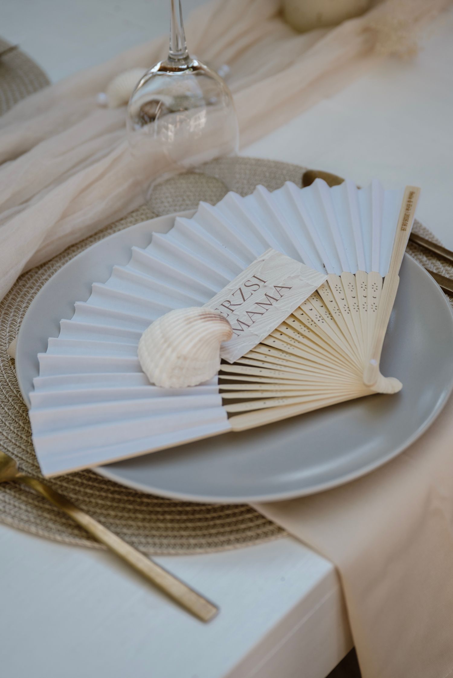 White paper fan and seashell on gray plate with gold cutlery at elegant wedding table setting.