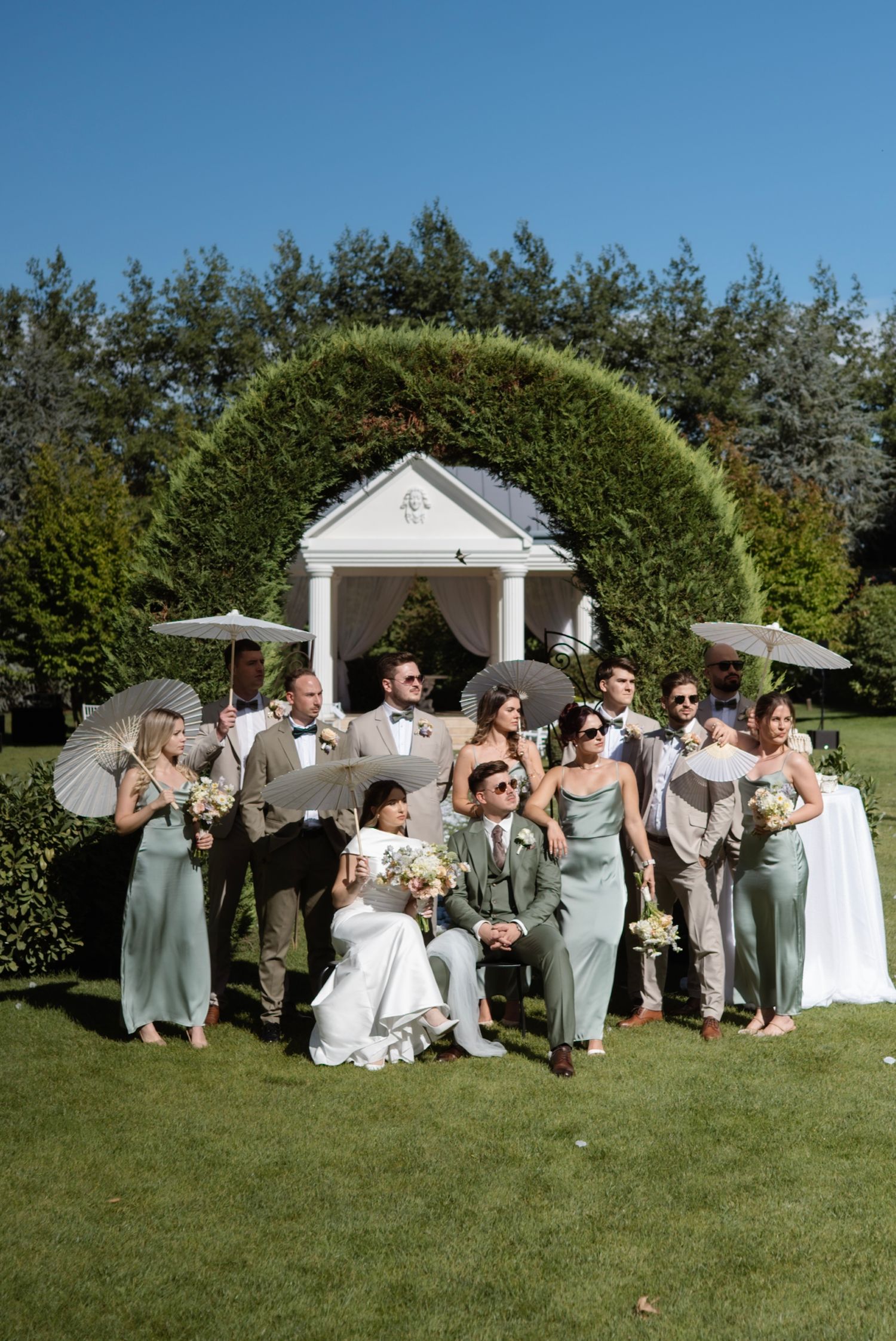Wedding party in sage green and tan attire posing outdoors with parasols near a white gazebo arch on a sunny day.