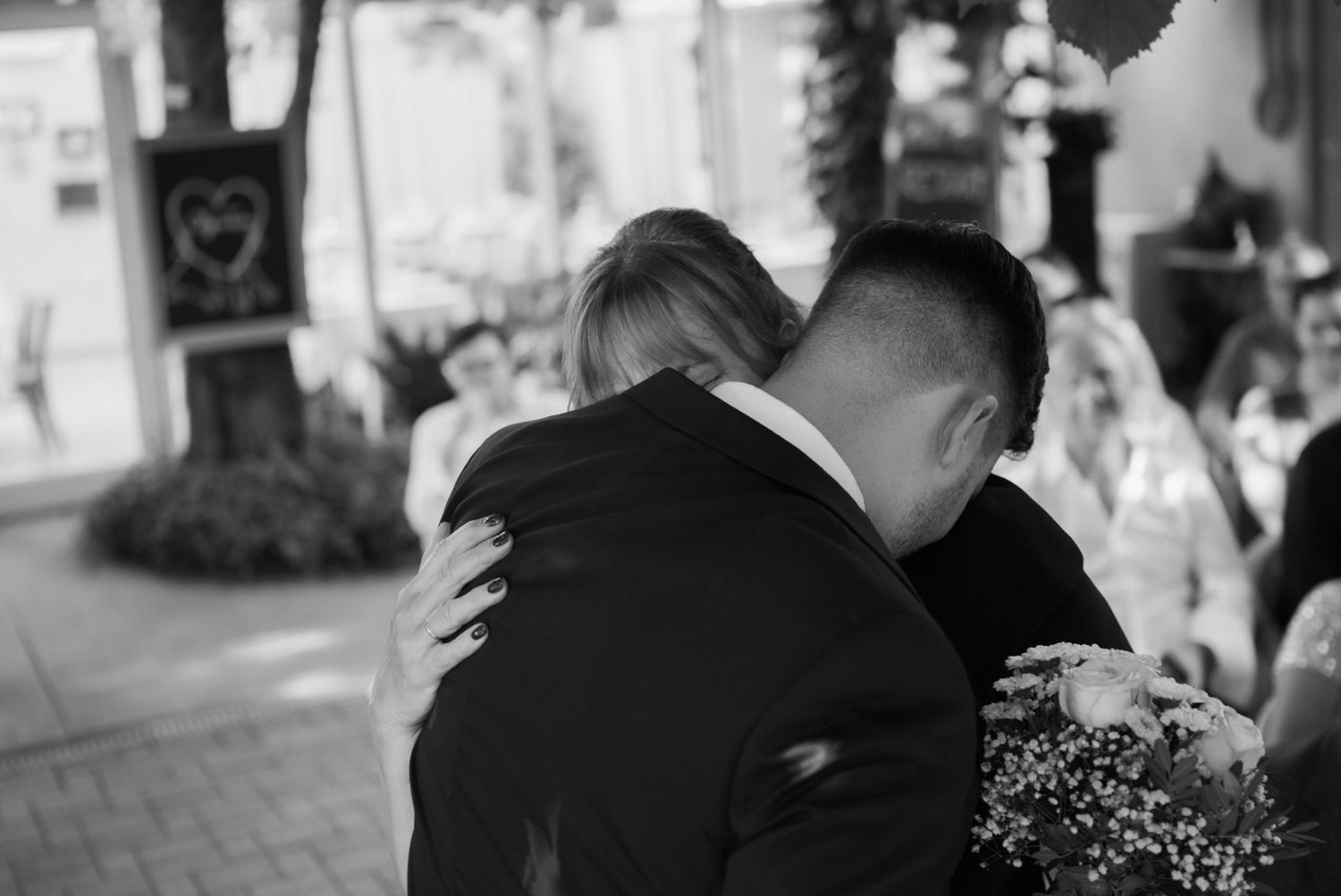 Black and white photo of a couple sharing an emotional wedding hug outdoors, with a heart sign visible in the background.