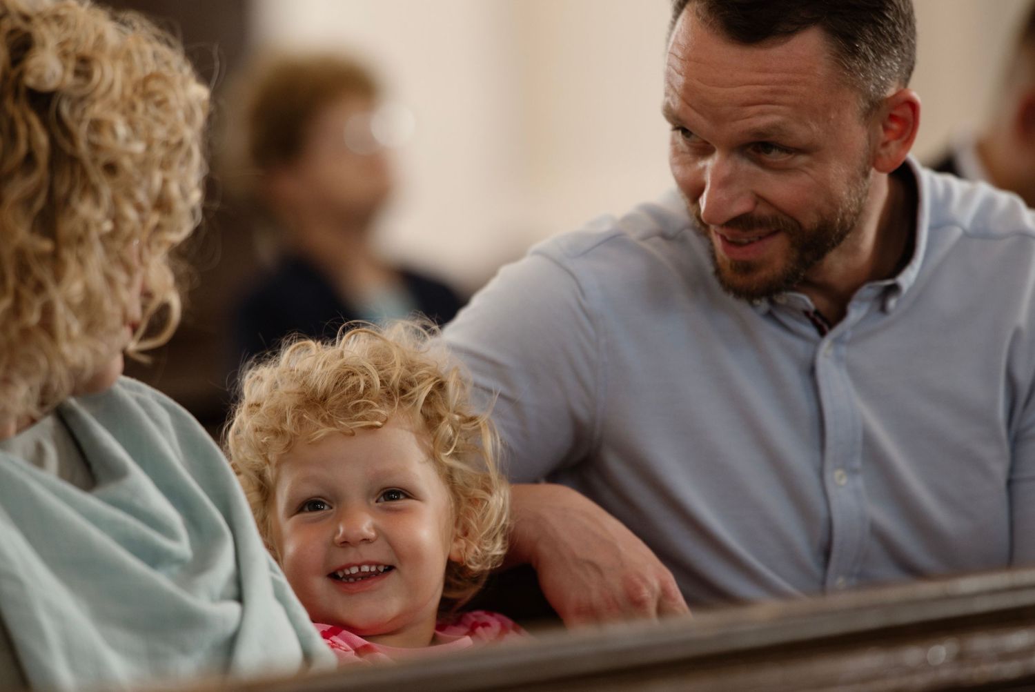 A smiling toddler with curly hair sits between two adults on a church pew, sharing a joyful family moment.