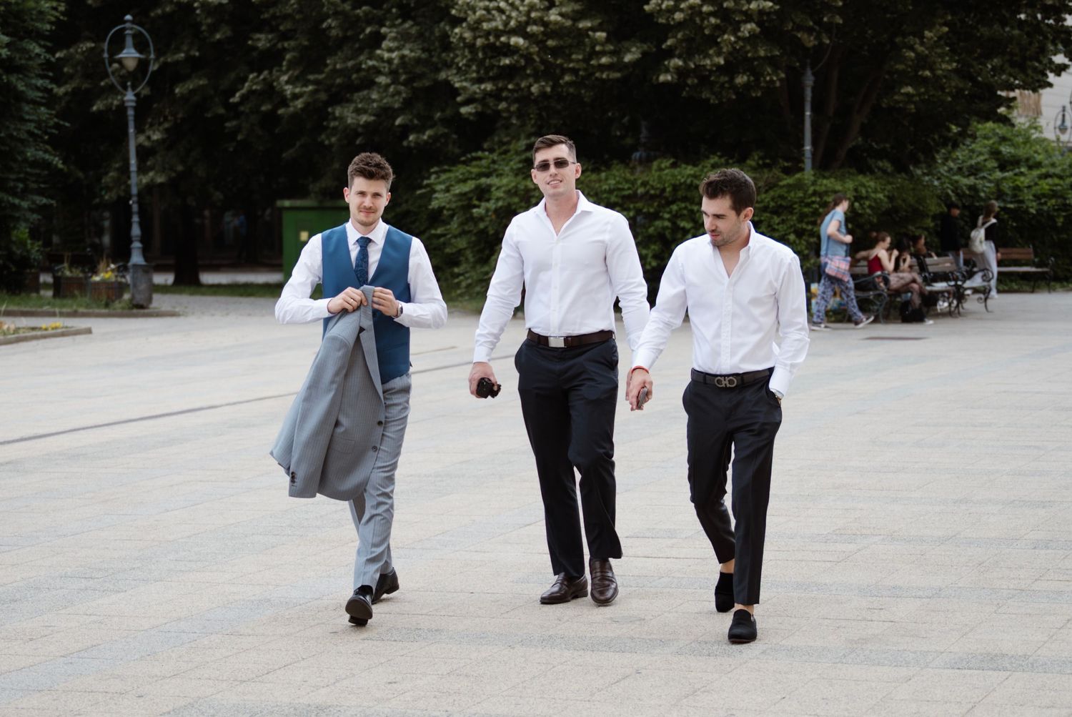 Three well-dressed young men in formal attire walking confidently across an outdoor plaza on a sunny day.