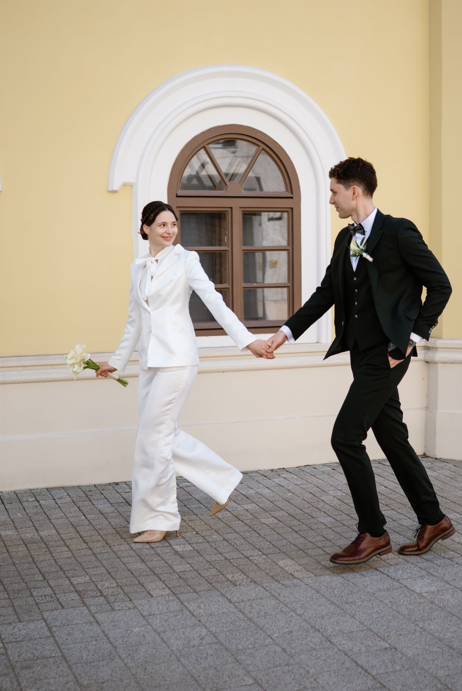 Bride in white pantsuit holds hands with groom in black suit, walking outside a yellow building on their wedding day.