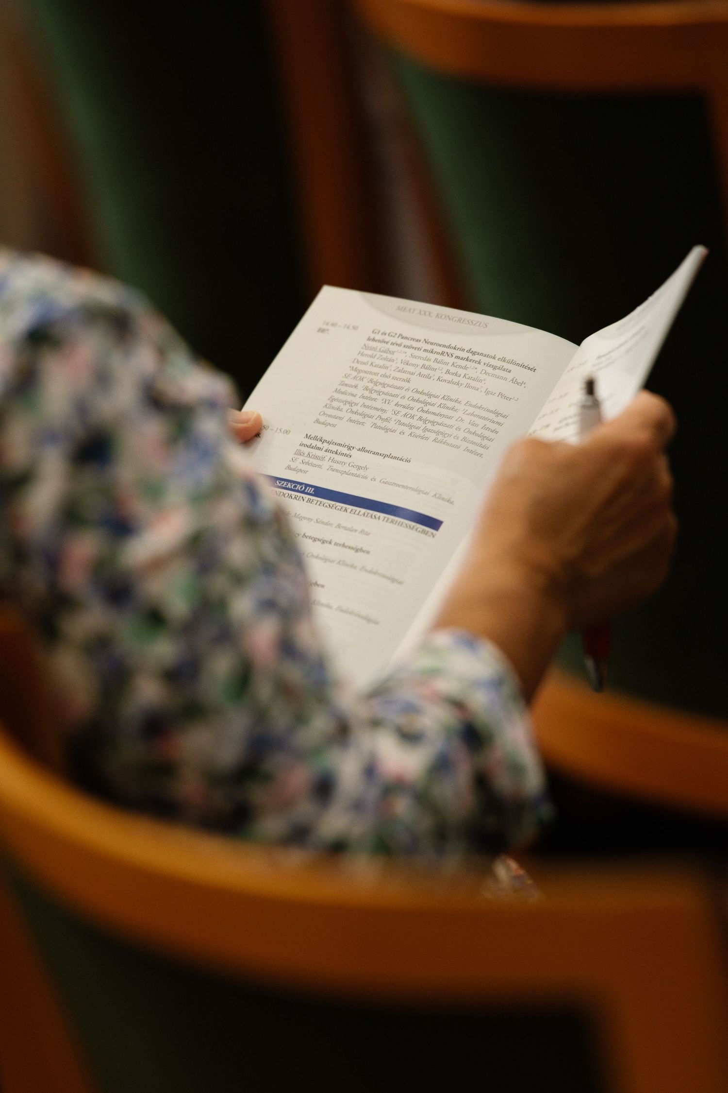 Someone reading a document or book while sitting in a chair with floral patterned upholstery in a dimly lit room.