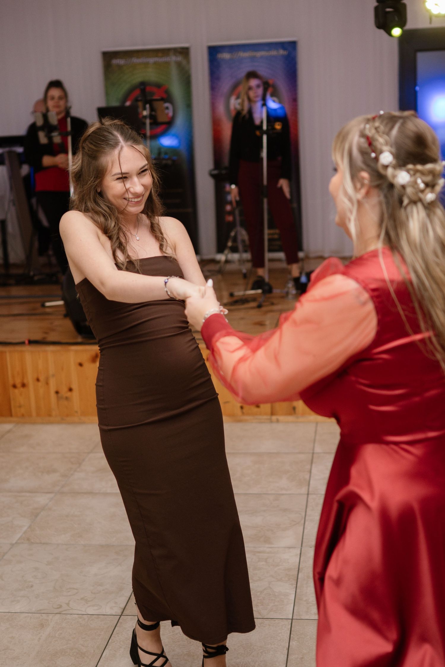 Two women dancing together at a wedding reception, one in brown and one in coral dresses.
