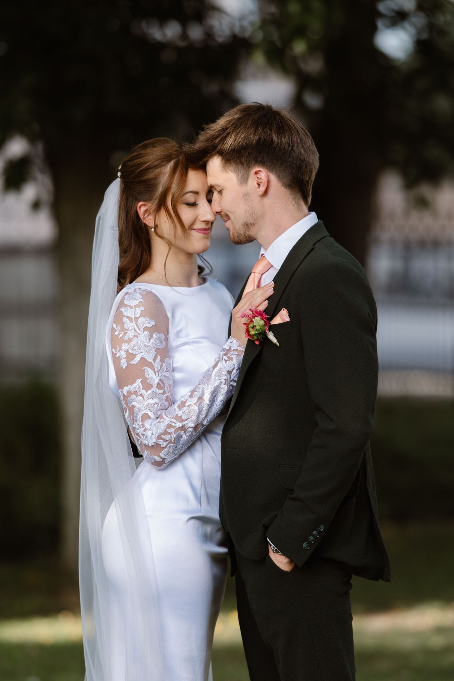 Romantic wedding portrait of couple in white lace dress and black suit embracing outdoors.