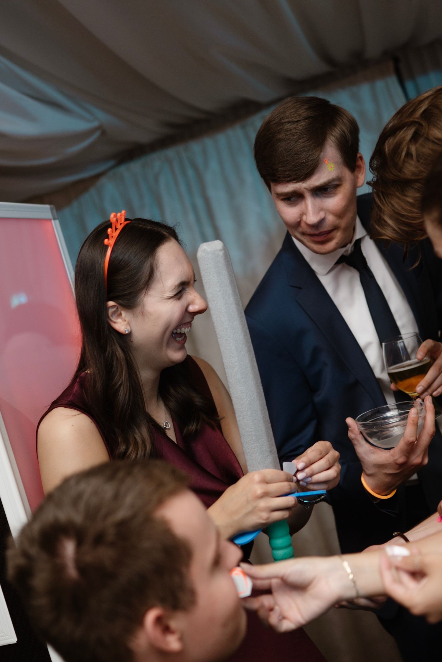 People laughing and socializing at a formal party, holding drinks and foam sticks at an indoor event.