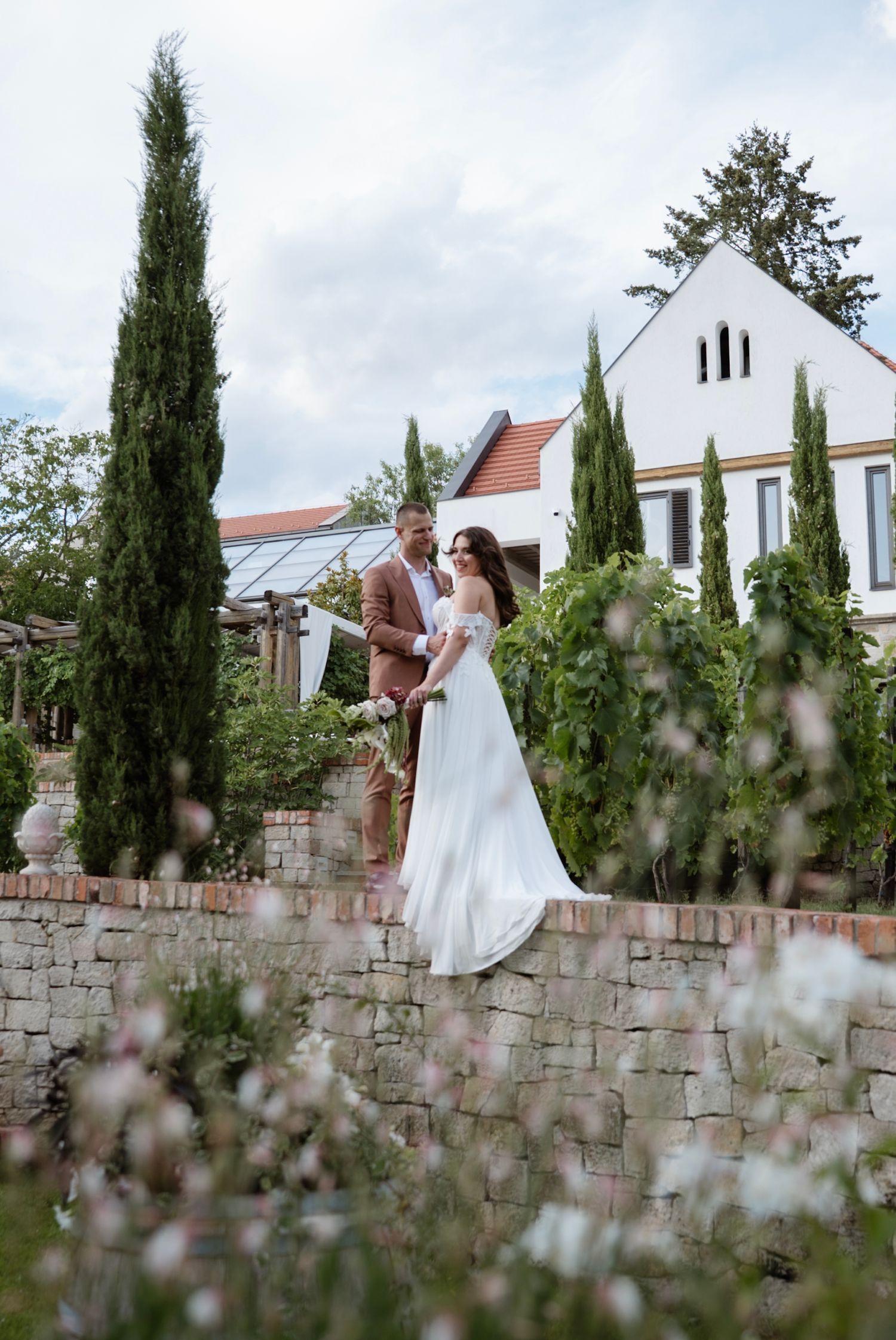 Bride and groom stand on stone garden wall surrounded by tall cypress trees at elegant estate wedding.