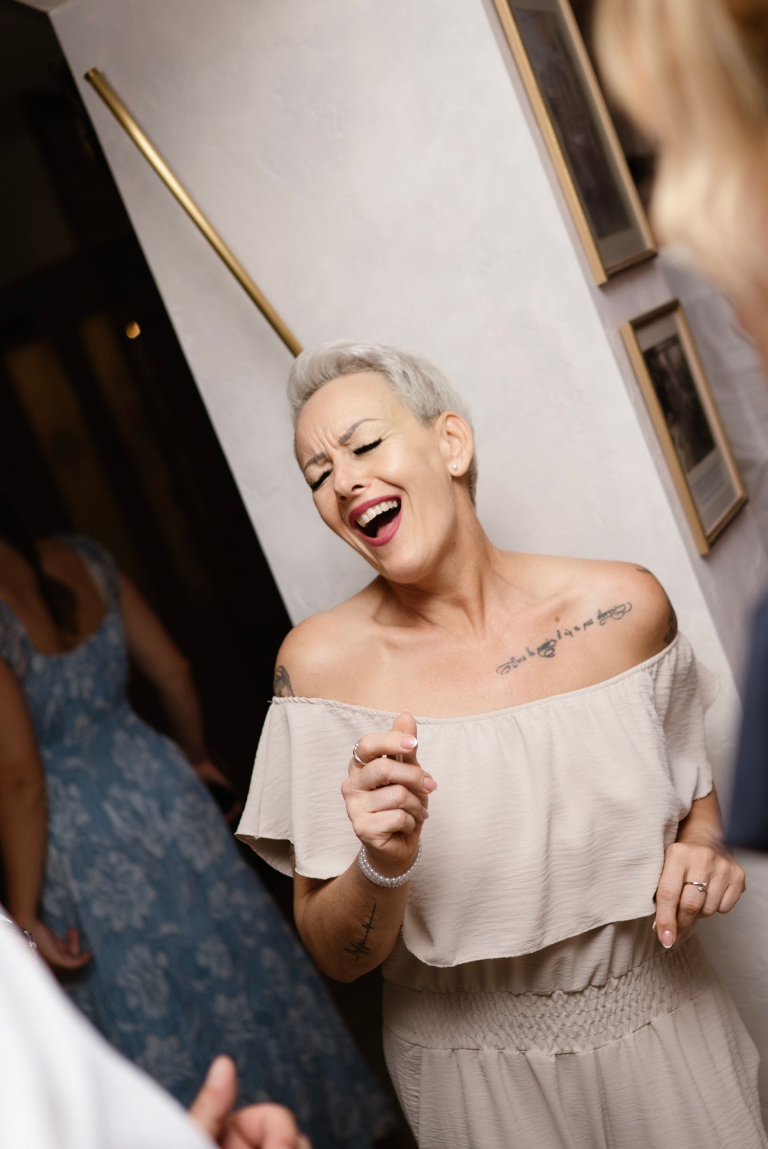 Woman with short gray hair laughing joyfully at a party, wearing an off-shoulder white dress with a chest tattoo.