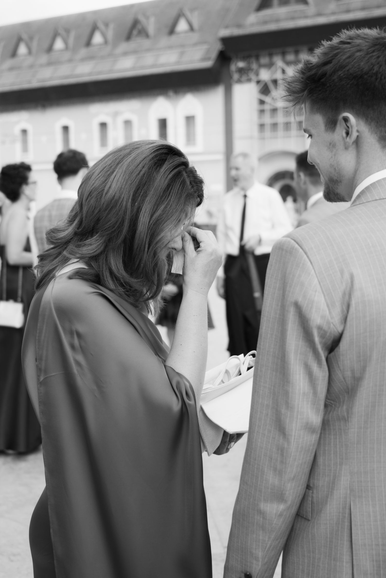 A woman wipes tears at an outdoor wedding ceremony while a man in a suit stands beside her.