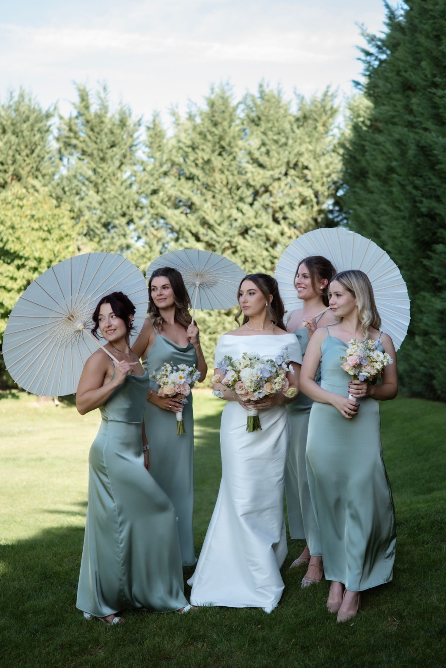 Bride in white gown with four bridesmaids in sage green dresses holding white parasols and bouquets outdoors.
