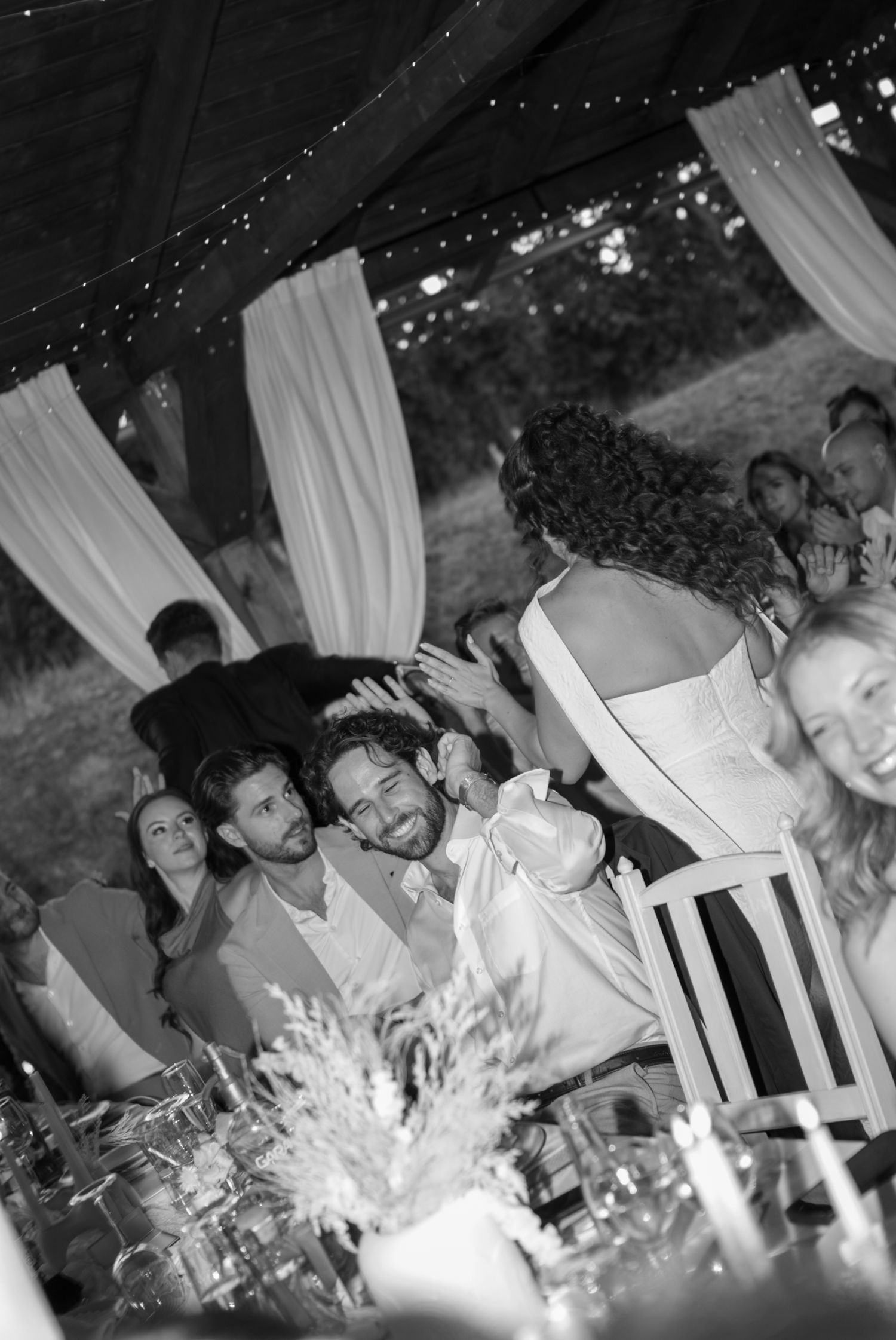 Wedding reception guests laugh and celebrate under draped tent with candlelit tables.