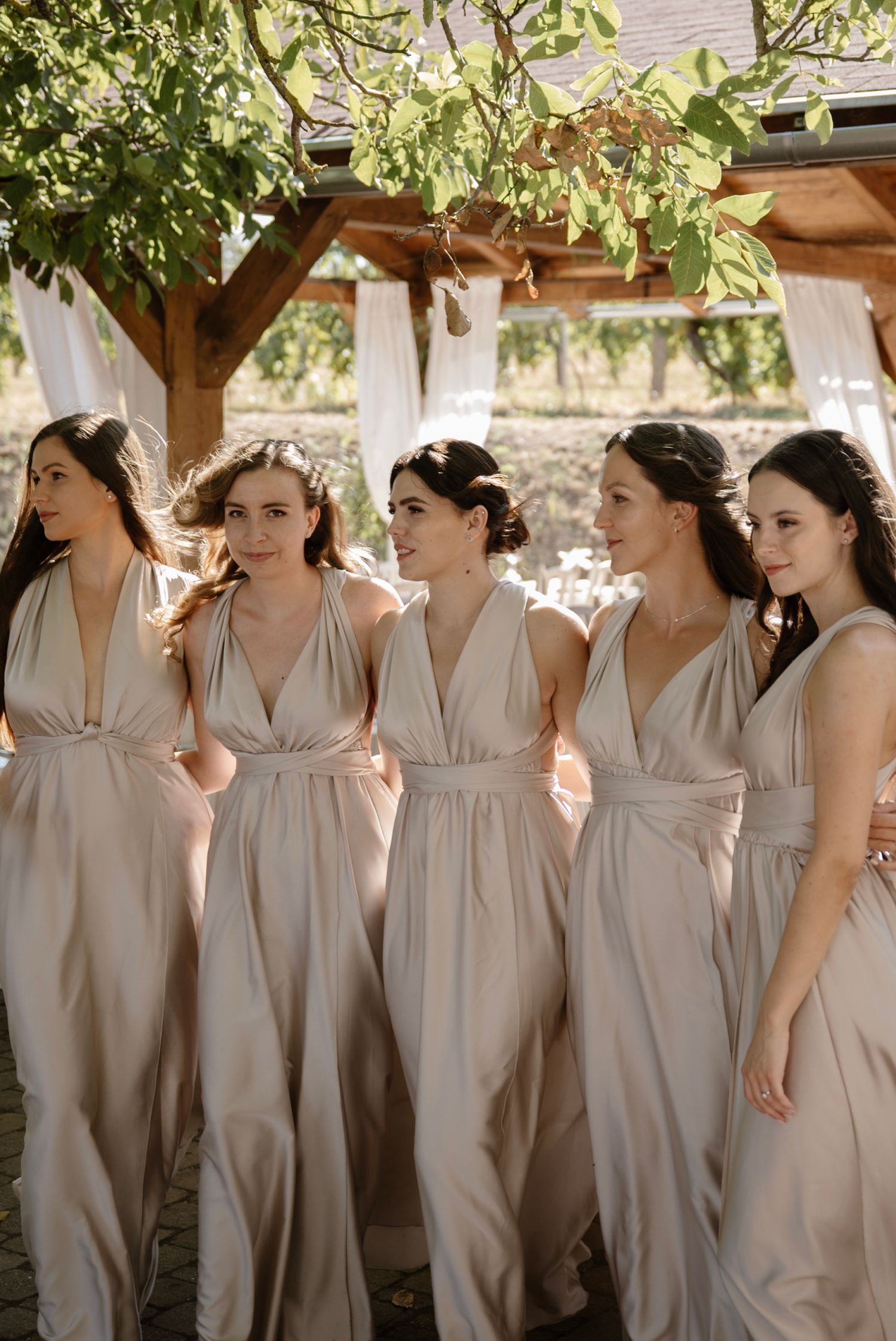 Five bridesmaids in matching champagne gowns walking together under a vineyard pergola.