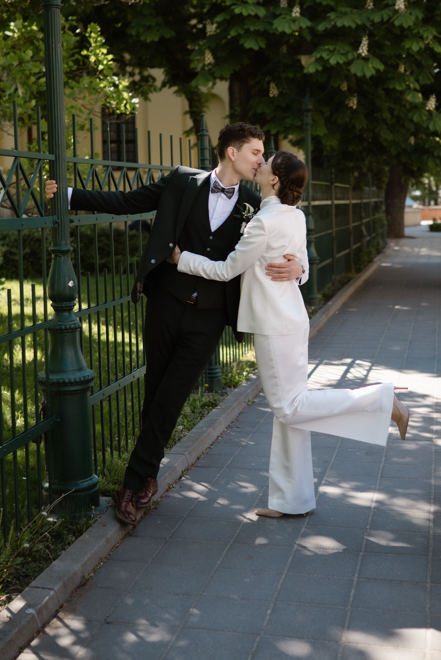 A couple in wedding attire shares a romantic kiss by an iron fence, surrounded by lush greenery.