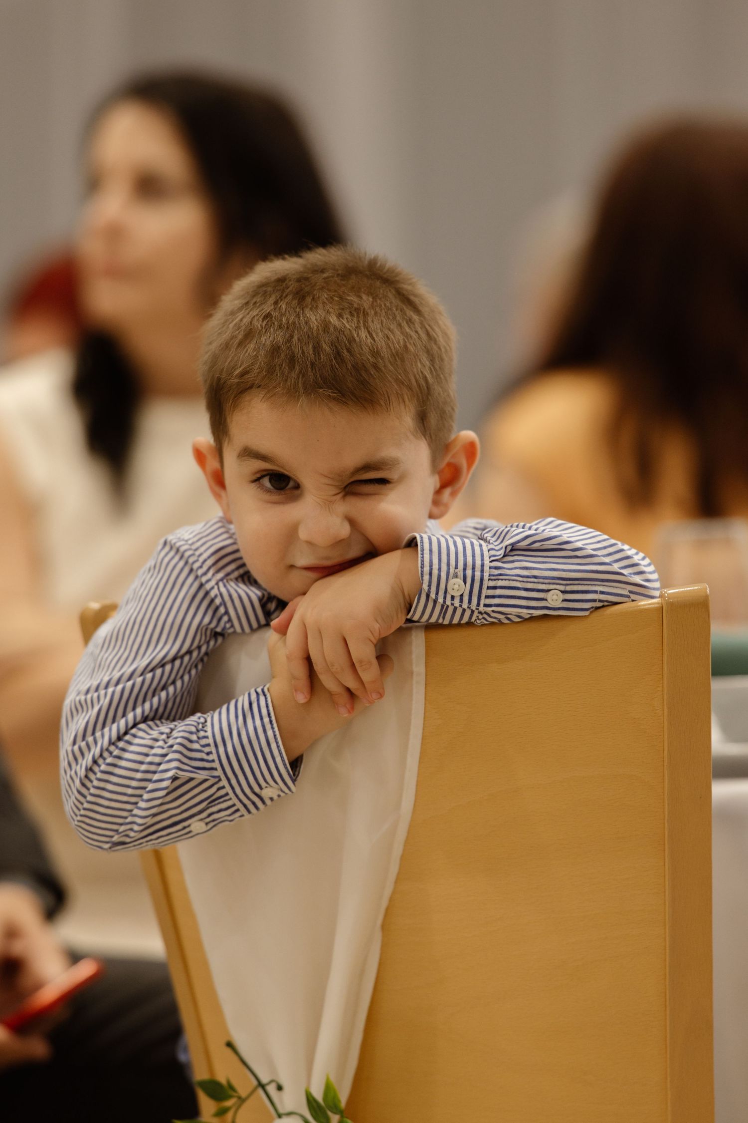 Young boy in striped shirt resting head on wooden chair back during formal event.