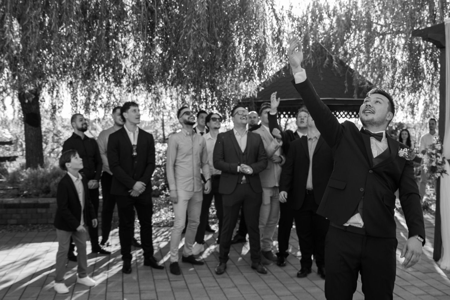 Wedding guests stand in a line outdoors beneath willow trees.