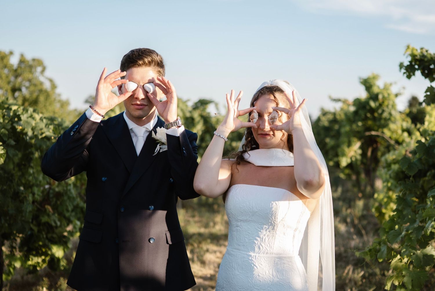 Bride and groom playfully hold wedding rings over their eyes in a sunny vineyard, wearing wedding attire.
