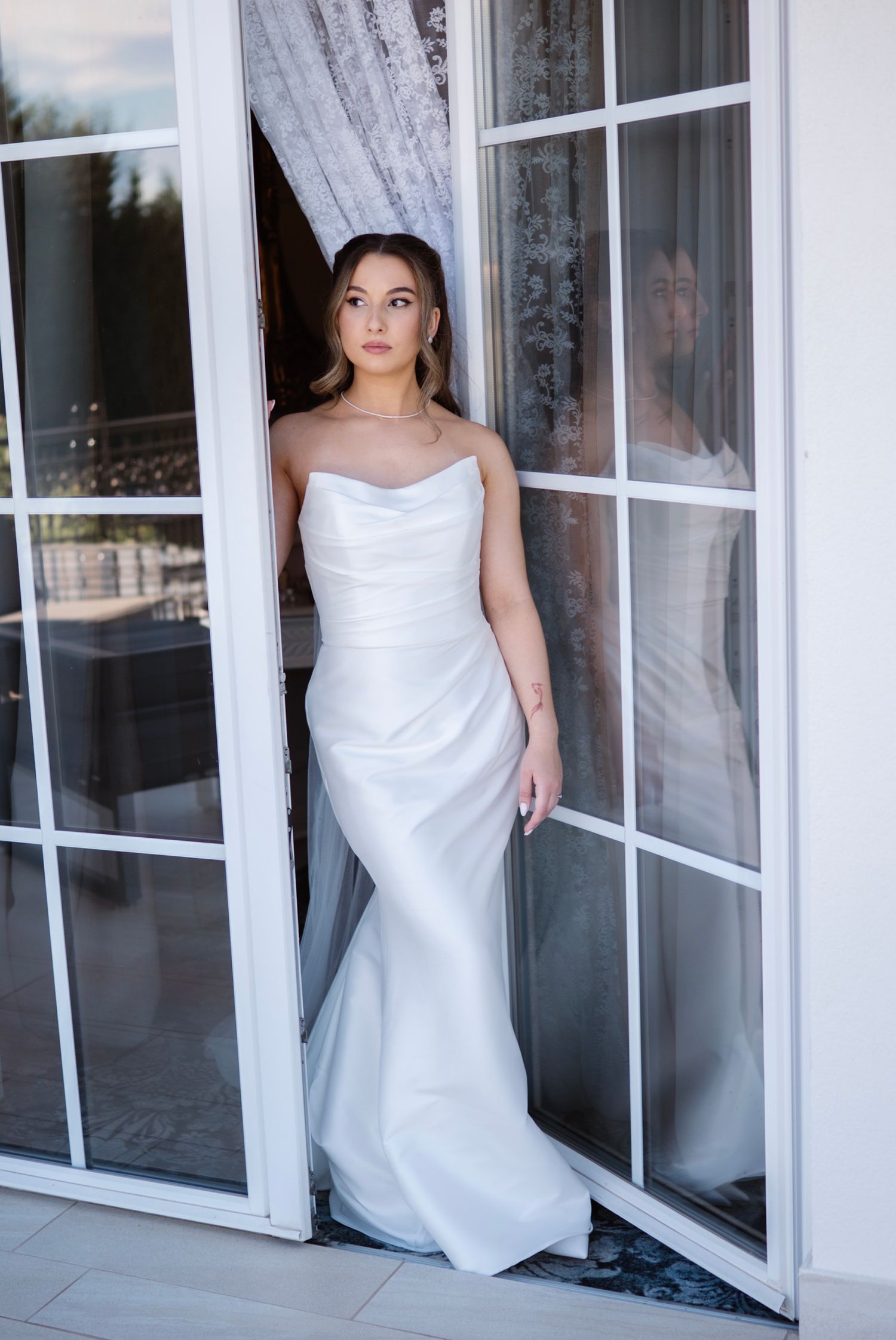 Elegant bride in a white satin sleeveless mermaid gown standing in French doors.